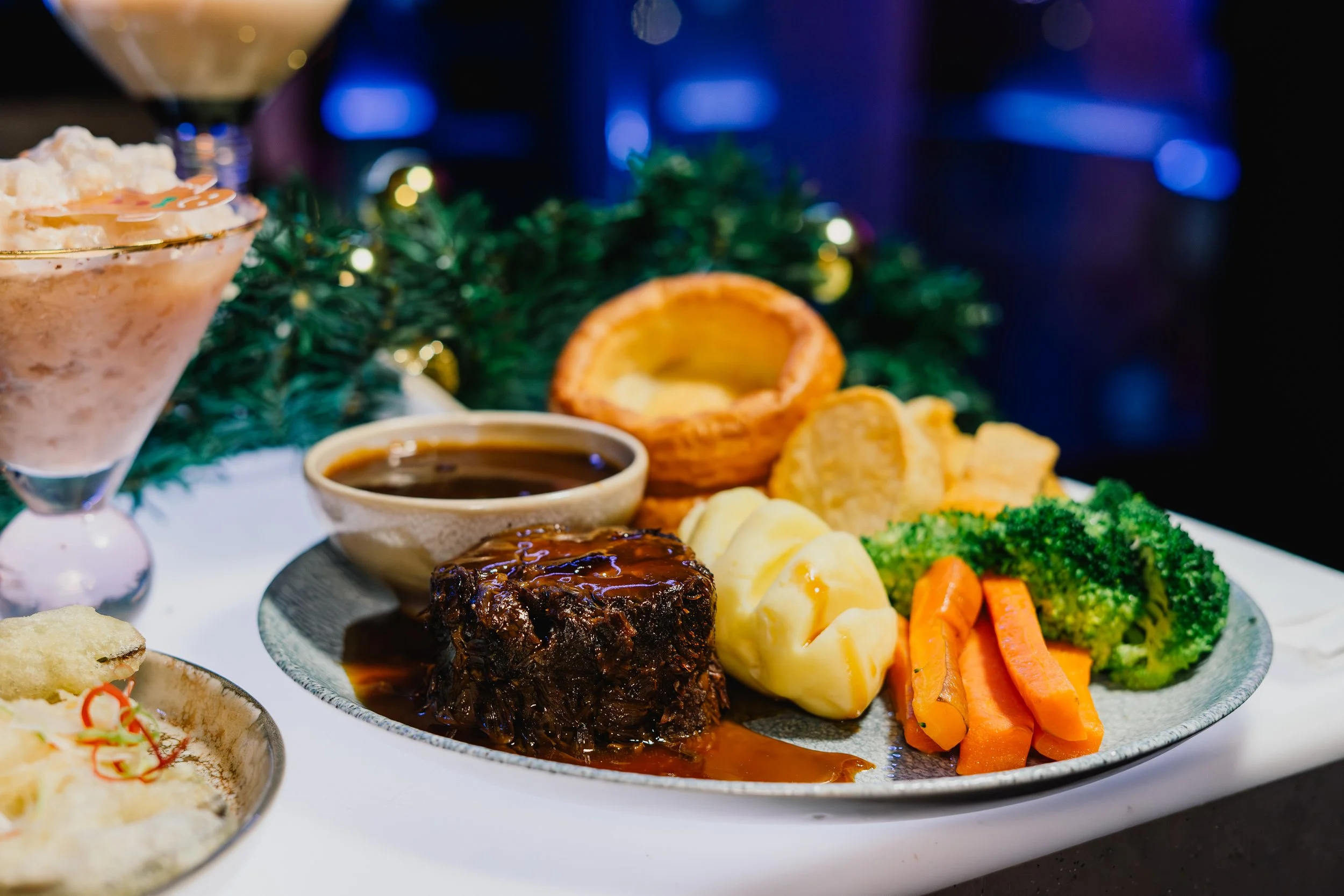 A festive meal featuring a roasted meat dish with dark gravy, surrounded by cooked vegetables including carrots, broccoli, and cauliflower, with a small bowl of gravy and a partially visible dessert or appetizer on the side. In the background, there 