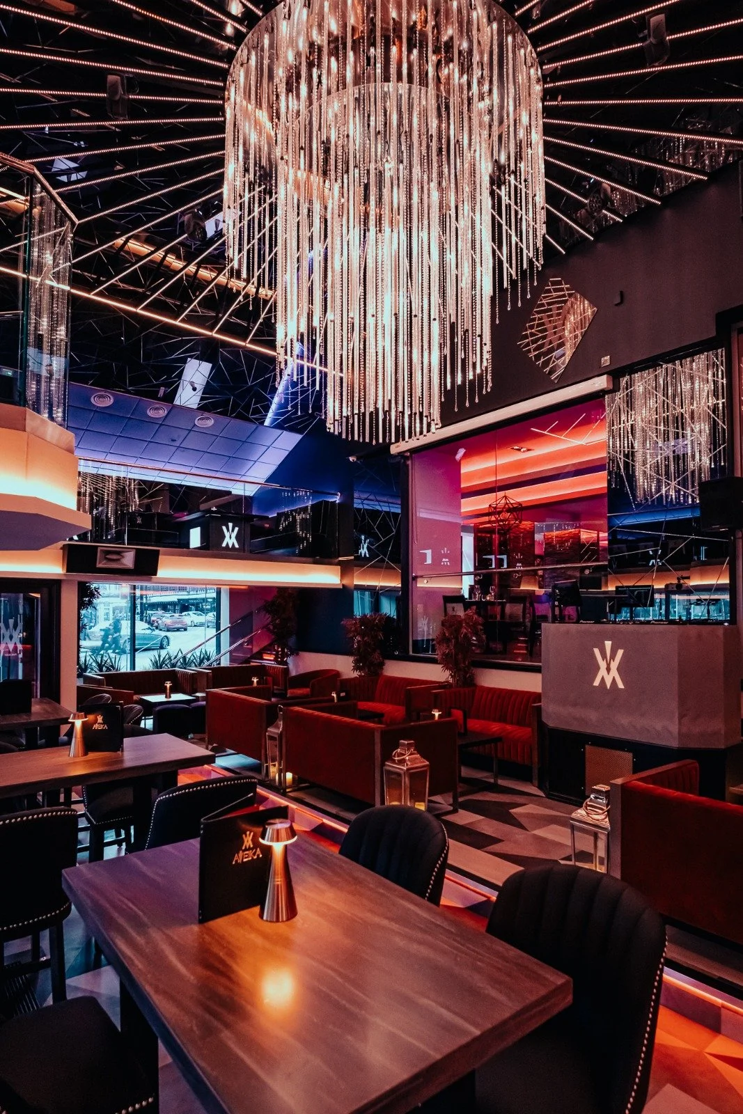 Interior of a modern lounge or bar with a large, circular chandelier with hanging lights, dark walls, red upholstered seating, and wooden tables.