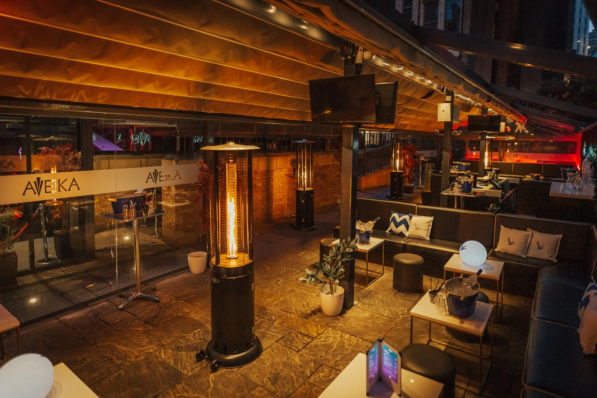 Nighttime outdoor patio with string lights, tall heaters, potted plants, and furniture, including a sofa with pillows and small tables.