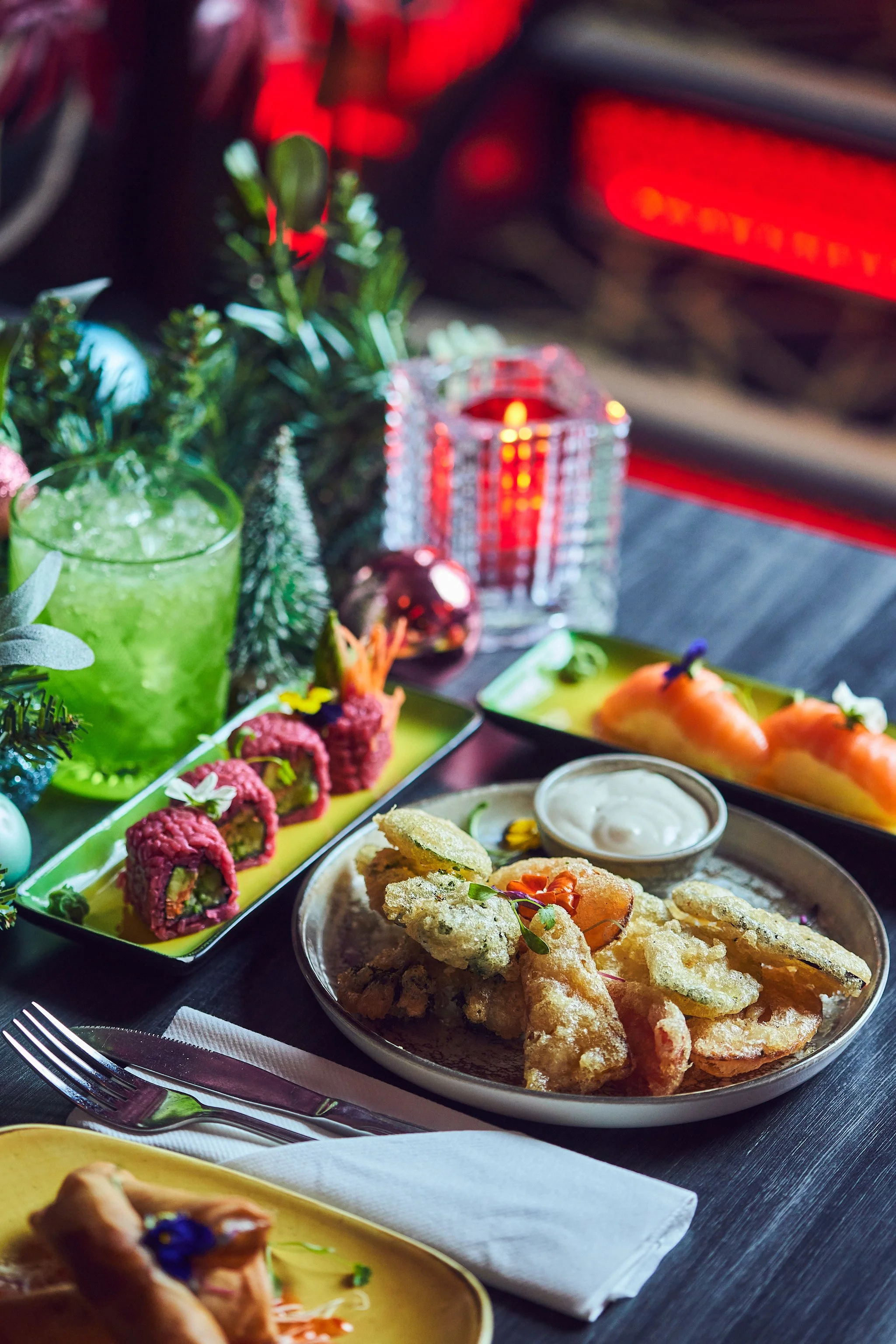 A variety of colorful dishes and drinks on a table, including sushi rolls, fried tempura, a green beverage, and small candles, decorated for a festive occasion.