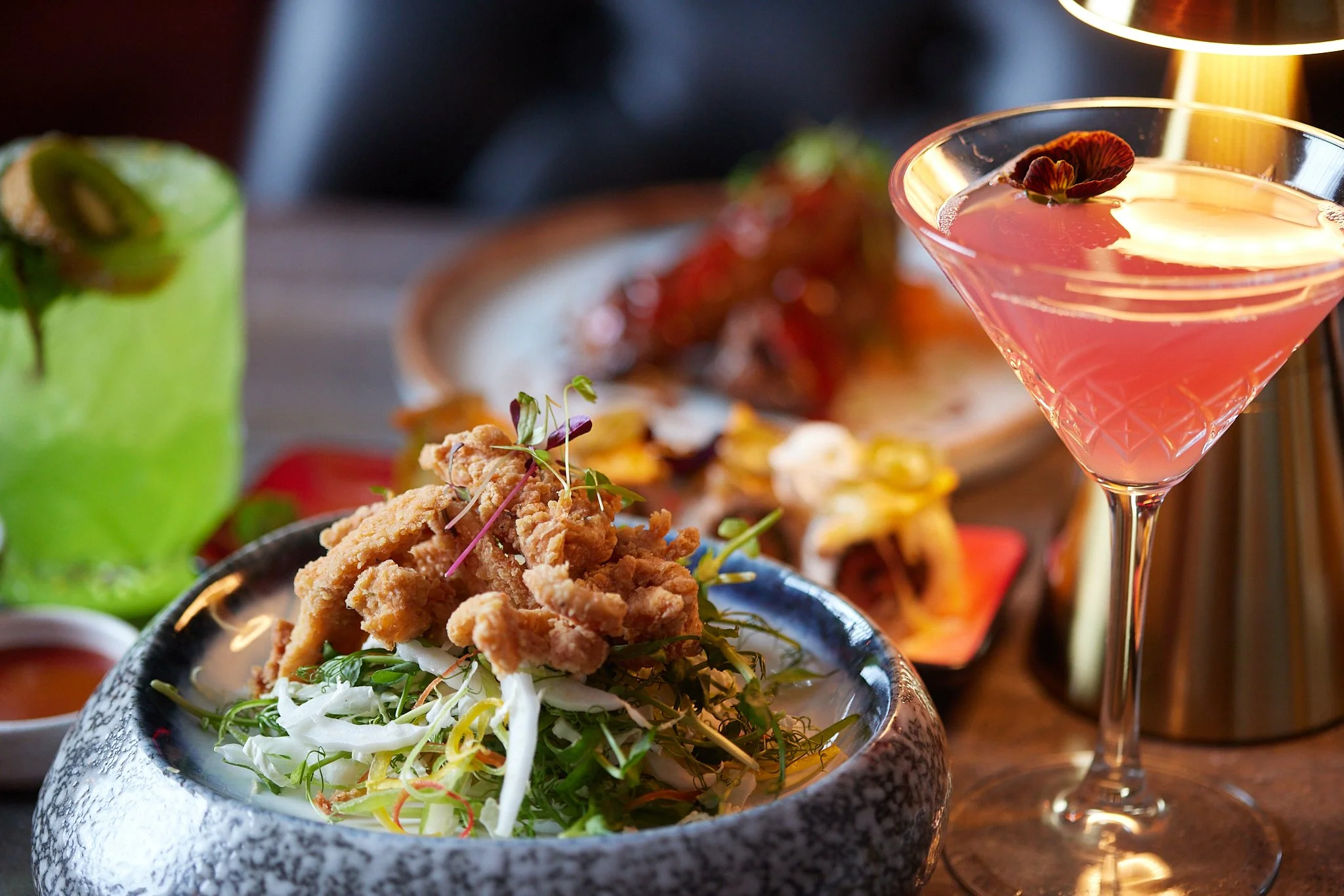 Close-up of a bowl of shredded chicken salad with greens, topped with microgreens, in front of a martini glass with a pink cocktail garnished with a flower petal. Other dishes and drinks are blurred in the background.