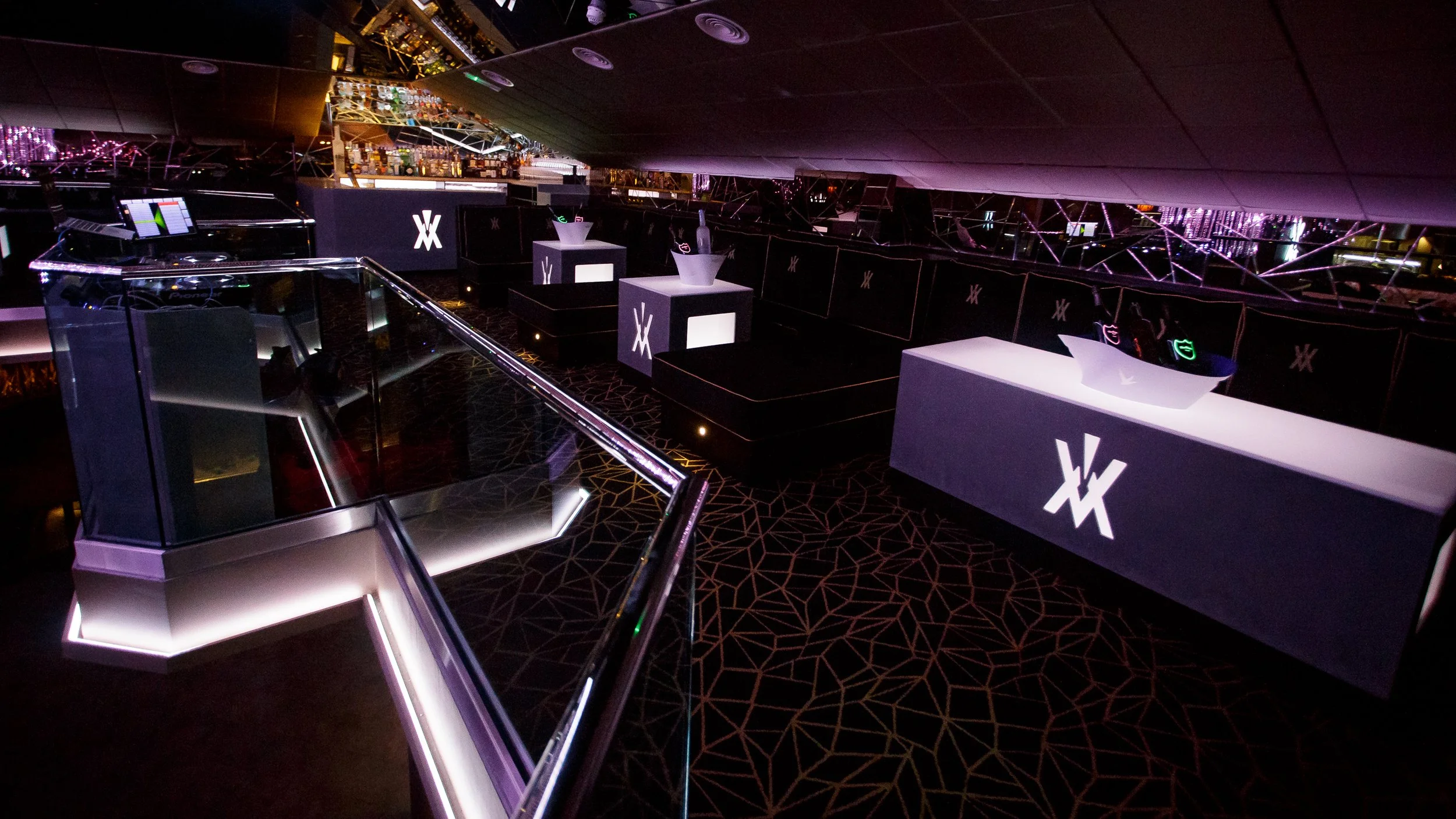 Interior of a modern nightclub with illuminated DJ booth, bar area, and seating featuring black and neon lighting decor.
