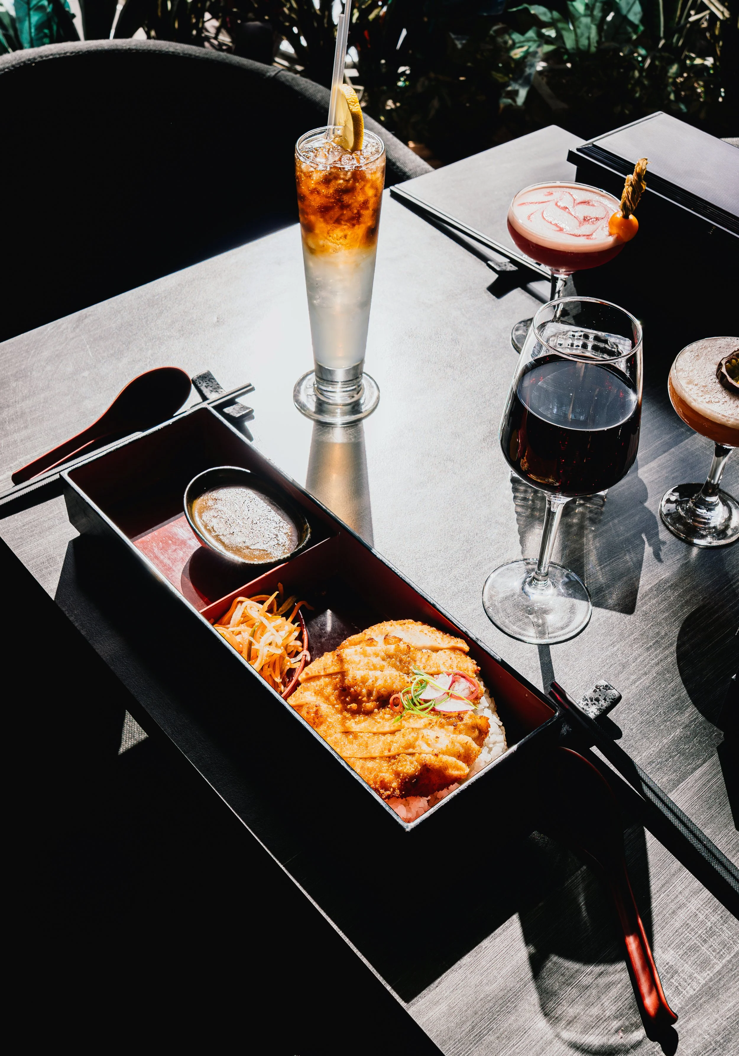 A table set with various drinks, including a tall cocktail with a lemon wedge and straw, a pink cocktail with a garnish, a glass of red wine, and a light-colored cocktail, alongside a Japanese bento box with fried fish, rice, salad, and dipping sauce