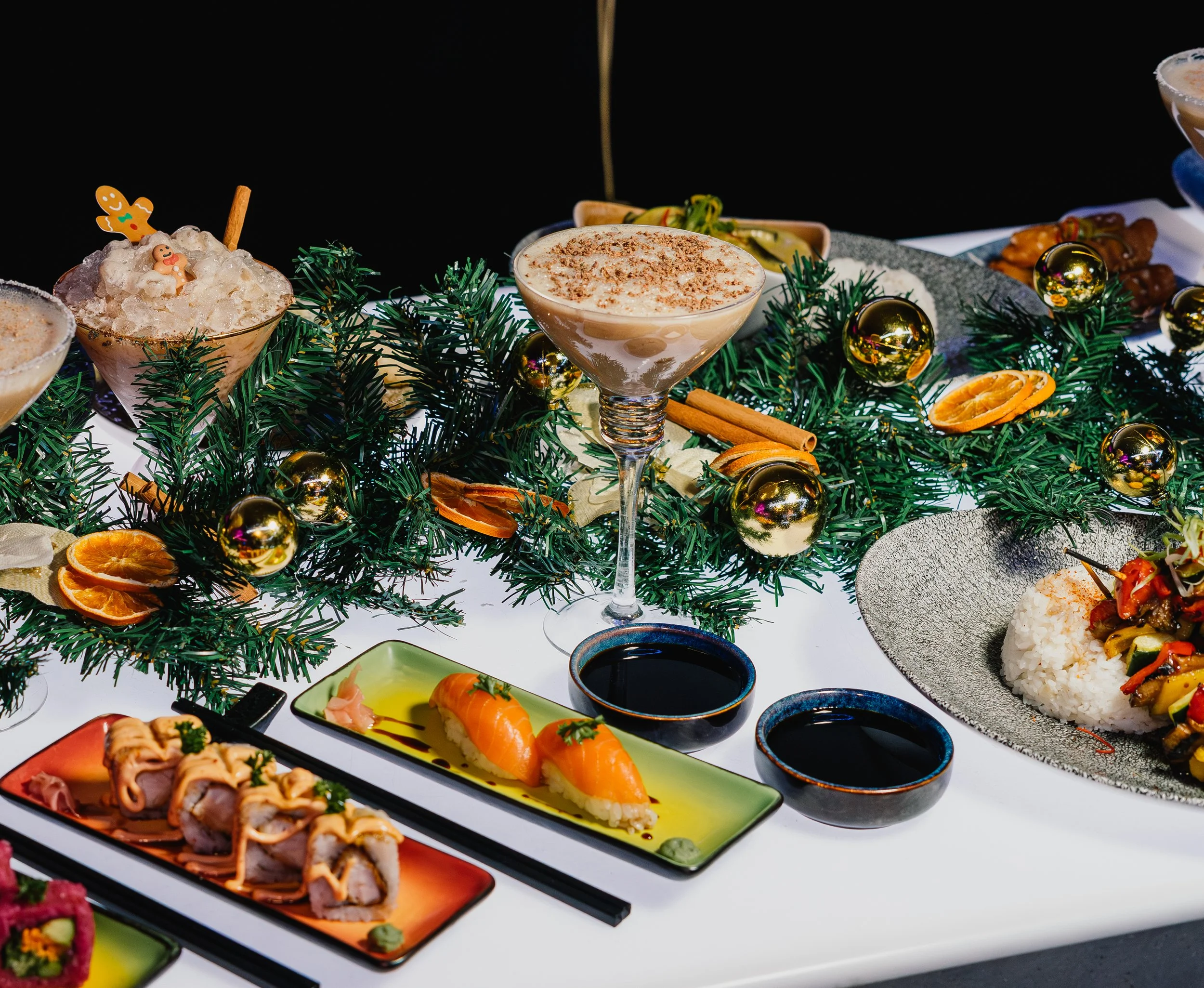 A variety of sushi dishes and desserts arranged on a table decorated with green pine branches and gold ornaments, with a black background.
