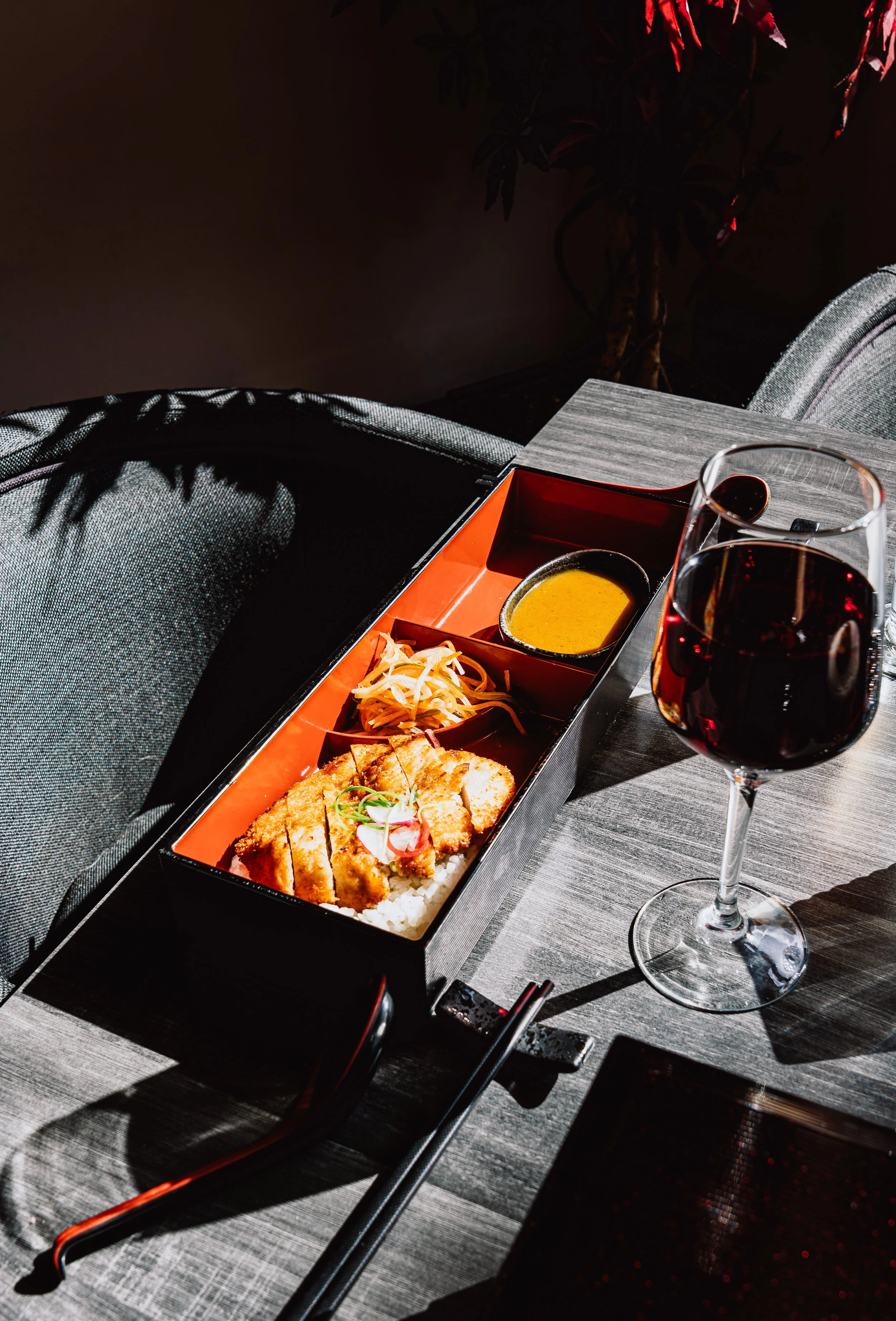 A black bento box with rice, fried chicken, salad, and sauce, alongside a glass of red wine, placed on a dark wooden table with a textured gray placemat, in a dimly lit setting with a potted plant in the background.