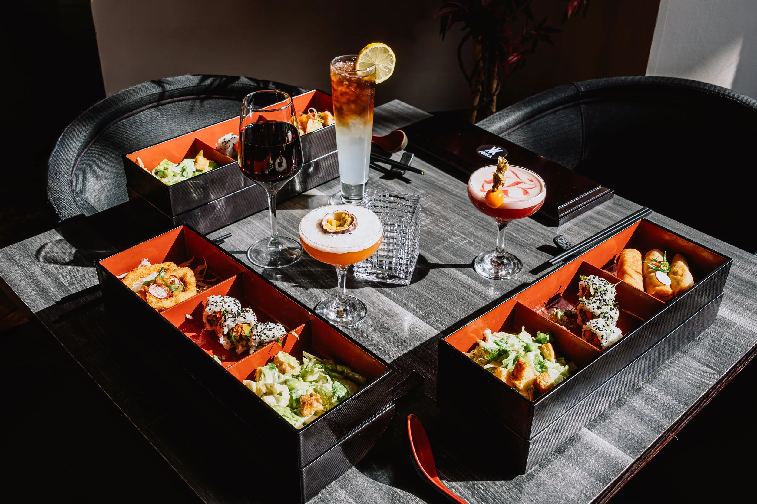 A table set with assorted Asian-style dishes in black and orange boxes, two glasses of wine, a cocktail with a lemon slice, and a glass of a layered drink with a garnish, all on a dark wooden table.