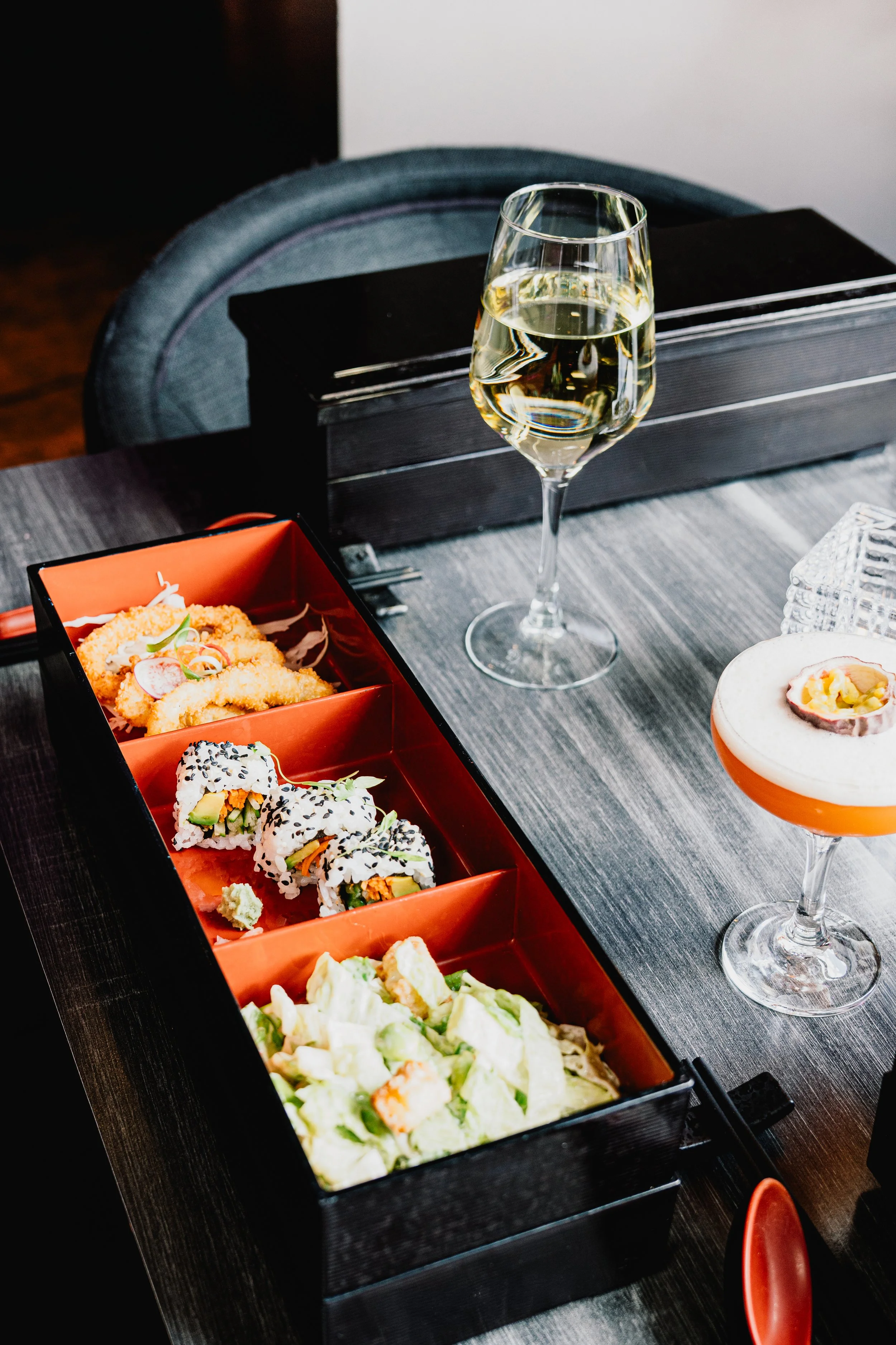 A black rectangular sushi box on a dark wood table containing different types of sushi rolls topped with sesame seeds. Two tall wine glasses filled with white wine and a bowl of salad are also on the table. A gray upholstered chair is in the background.