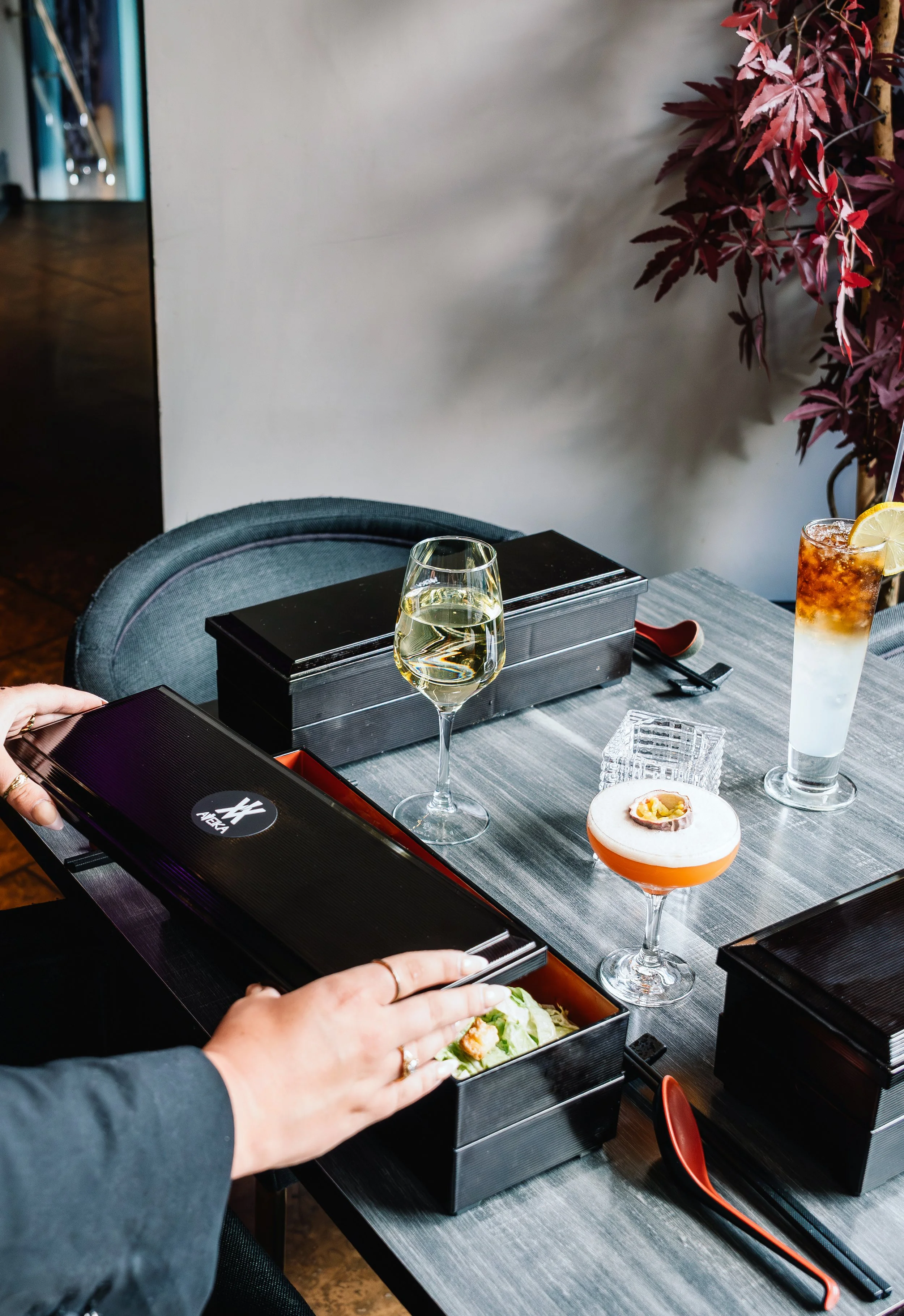 A dining table with two black boxes, one containing a salad and the other a beverage. A glass of white wine and a cocktail with a lemon slice are also on the table. A person is reaching into the salad box. The background has a minimalistic wall and a