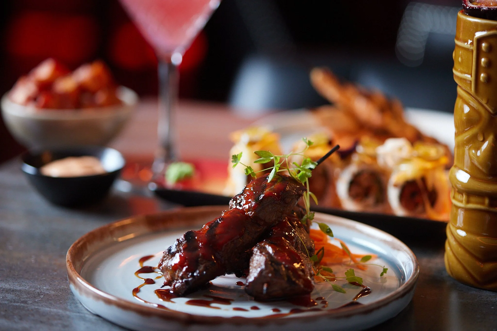 A glazed meat rib garnished with herbs on a white plate, with a background of blurred dishes and a dark setting.