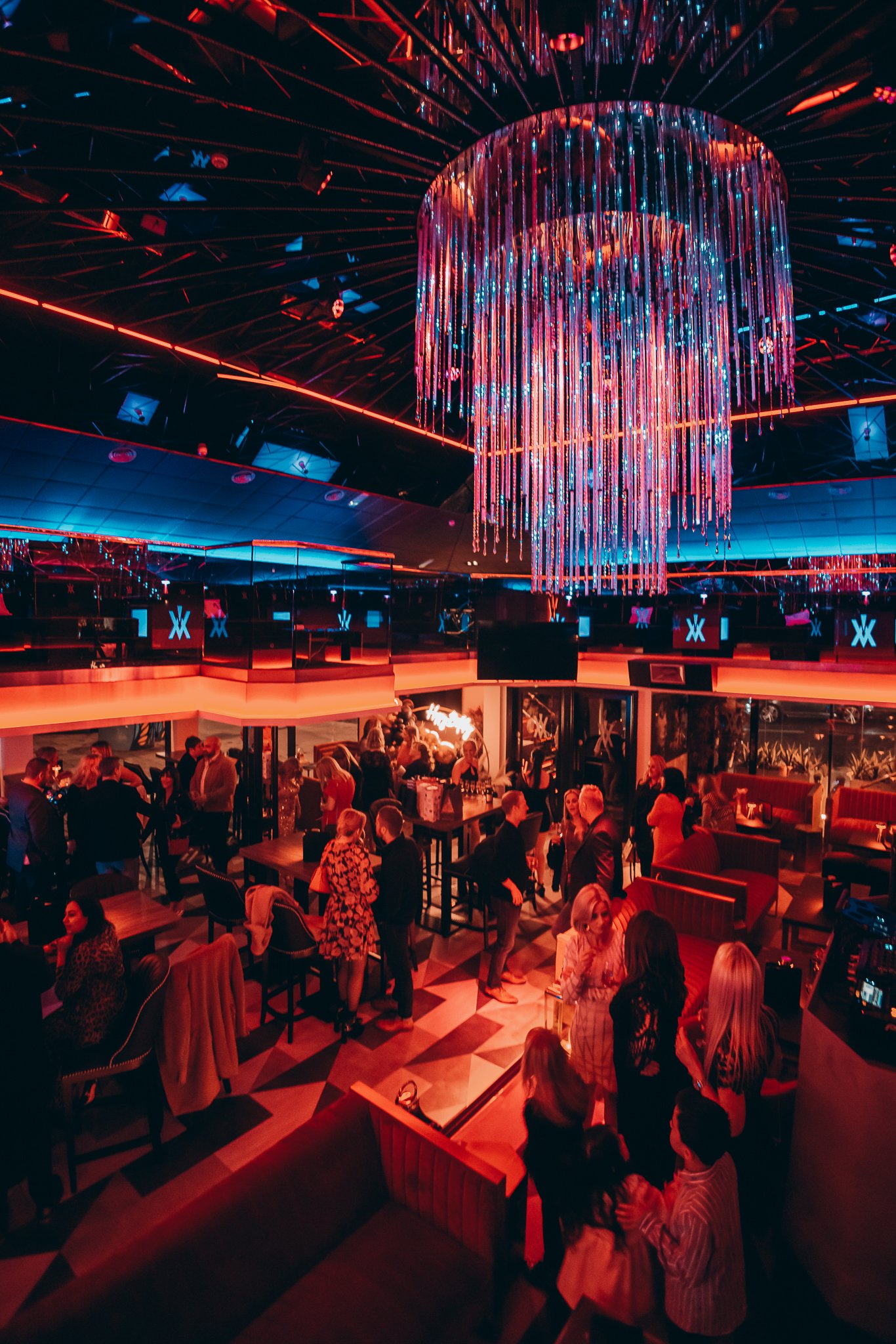 A lively nightclub scene with a large, illuminated chandelier hanging from the ceiling. The club is filled with people sitting and standing, enjoying drinks and socializing. Red and blue lighting accents surround the space, creating an energetic atmo