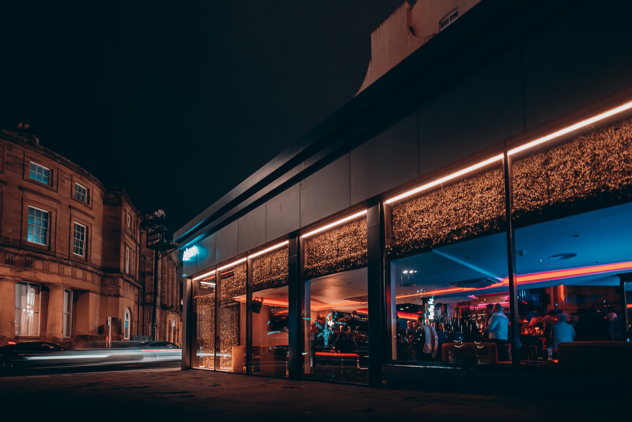 Night view of a modern building with neon lighting and glass facade reflecting city lights, with vintage-style brick buildings in the background.