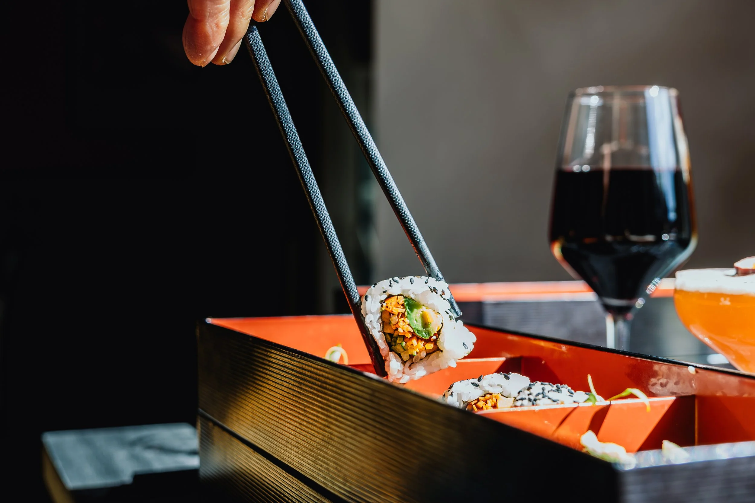 Close-up of a sushi roll being picked up with black chopsticks, with a glass of red wine and a glass of amber-colored beverage in the background on an orange tray.