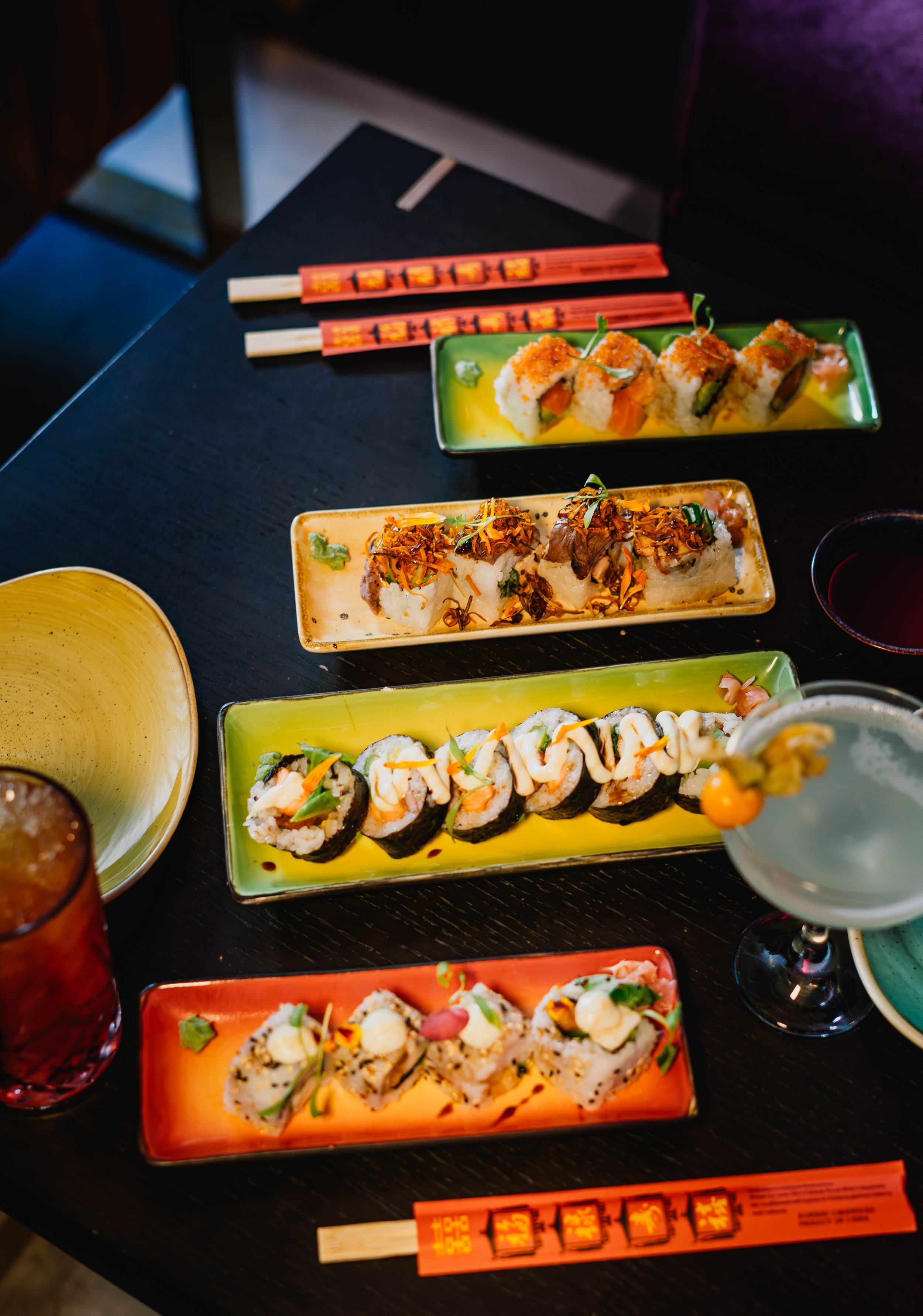 Assorted sushi rolls on rectangular plates, chopsticks, a drink, and a bowl on a dark table.