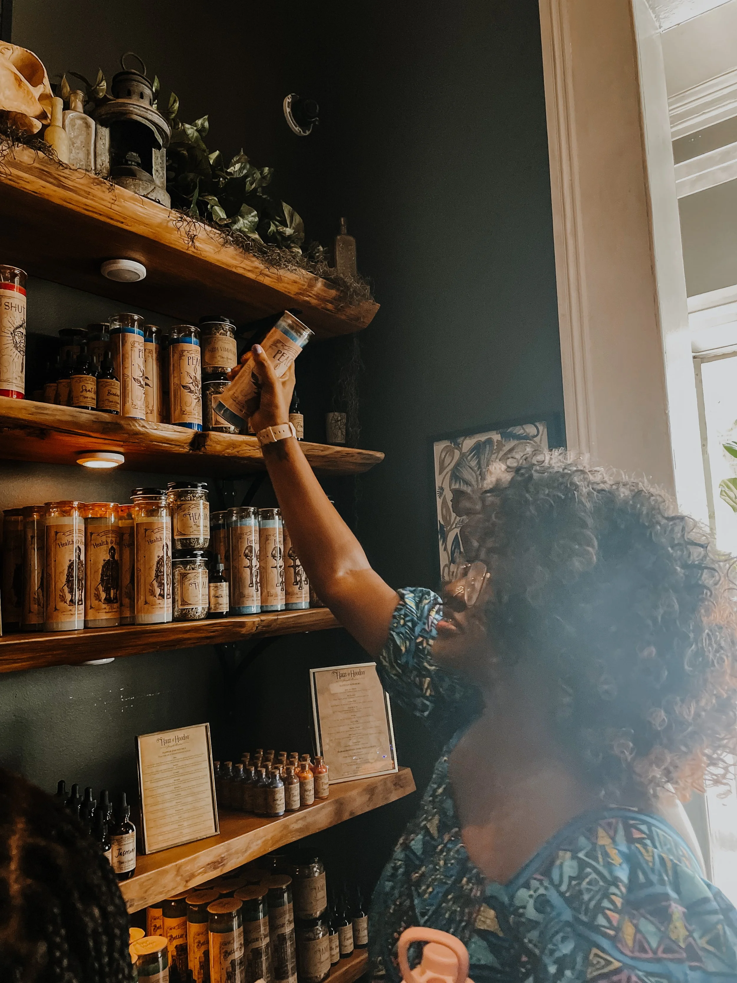 A woman with curly hair browsing products on a wooden shelf inside a store. The shelf contains various bottles and containers, some labeled 'Peace' and 'Health'. There is natural light coming from a window on the right side.
