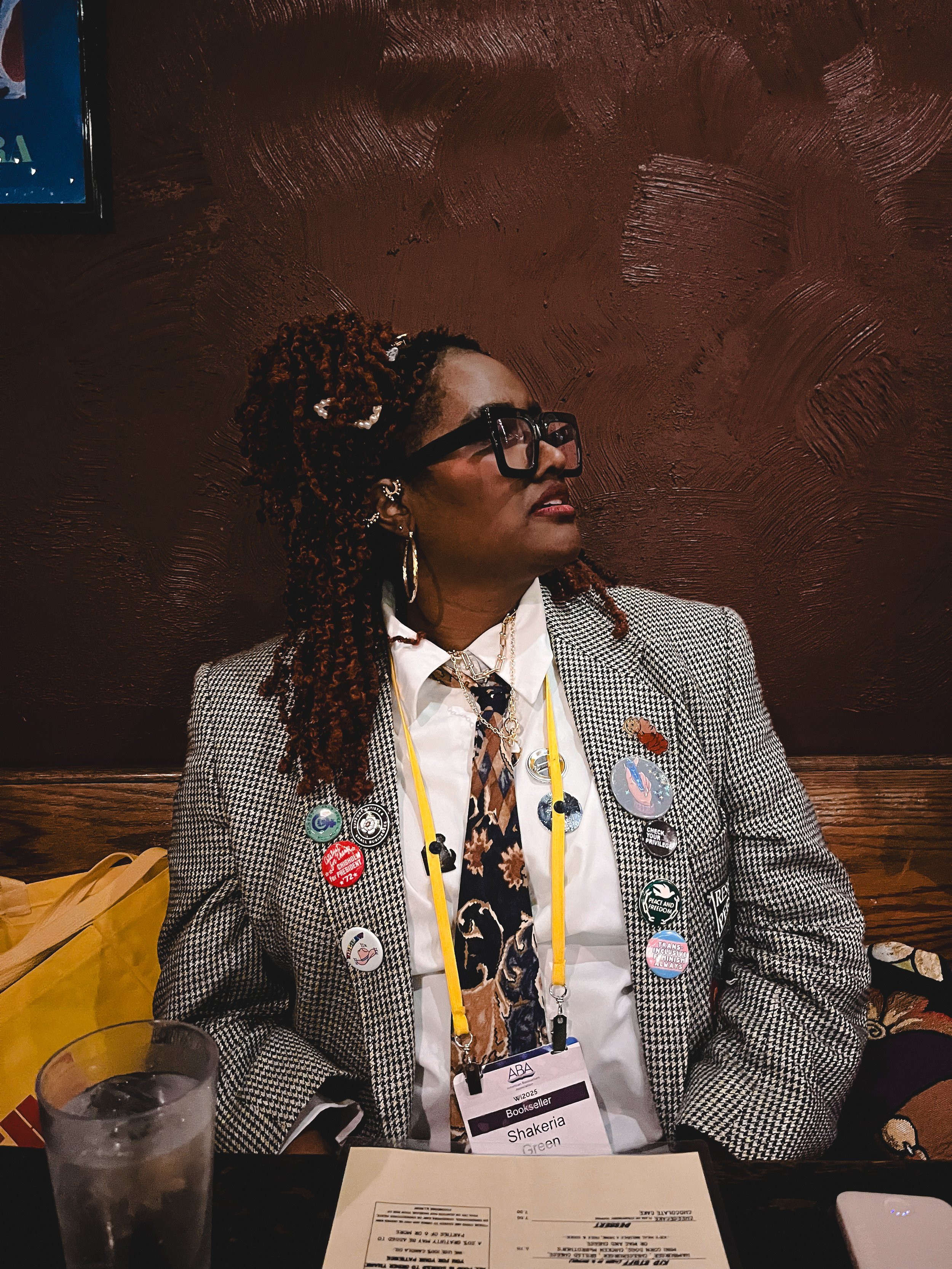 A woman with curly hair, glasses, and multiple buttons on her jacket, sitting at a table with a drink and papers, in a dark room with a textured brown wall and a yellow bag on the side.
