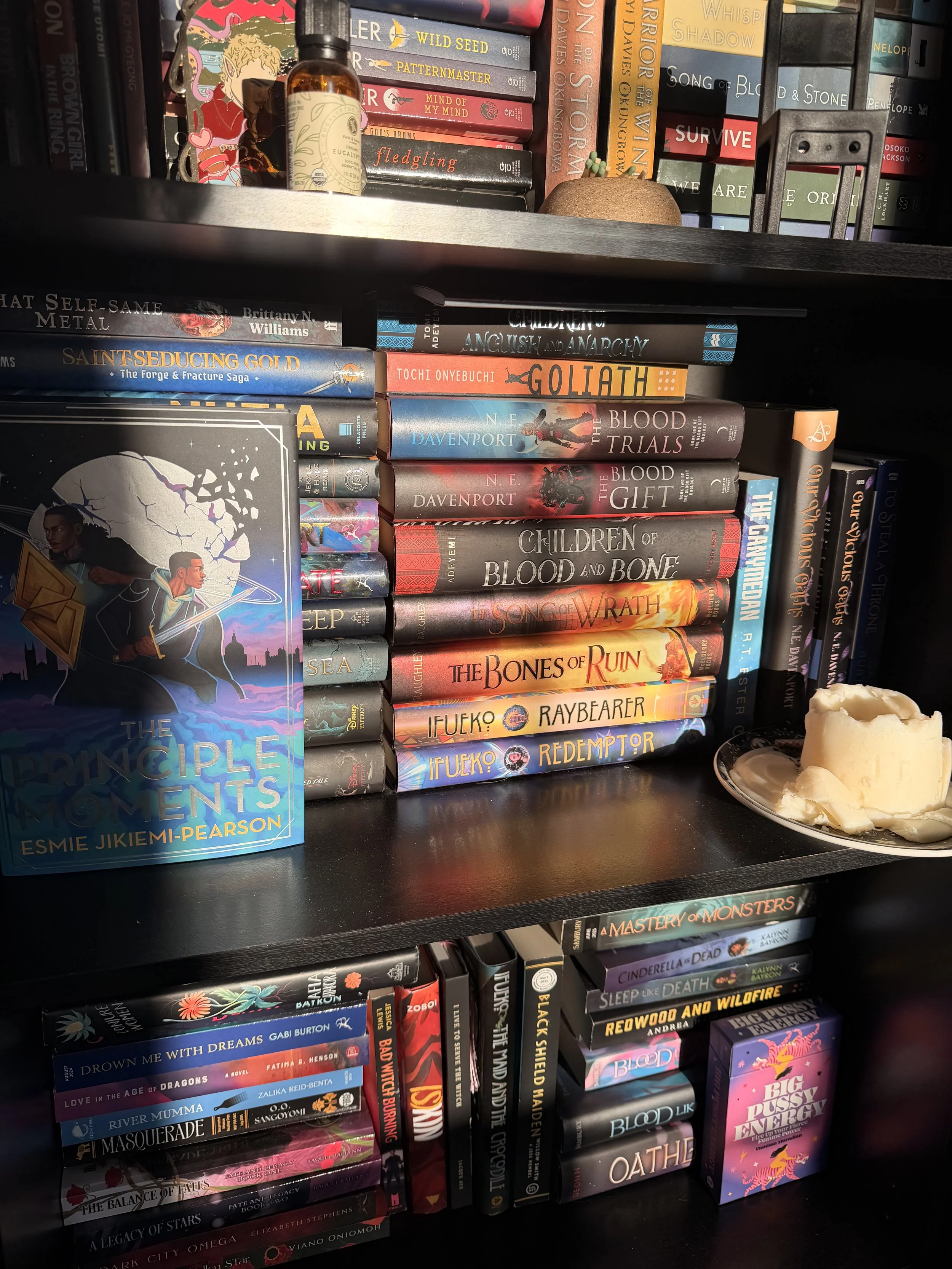 A bookshelf filled with a variety of colorful books, some stacked horizontally and others vertically, with a small bottle and a decorative item on the top shelf. There is a candle with melted wax on a plate in the foreground.