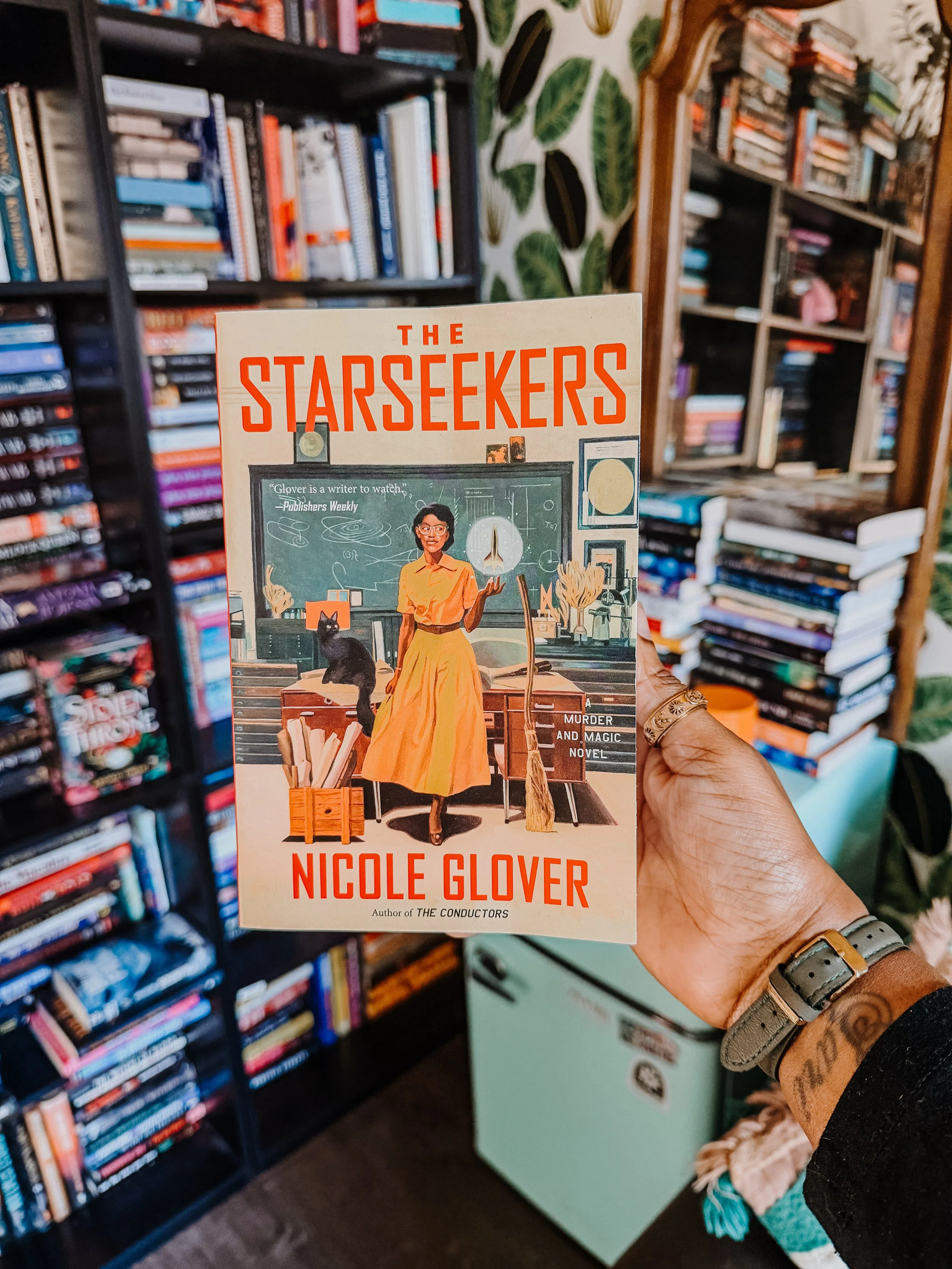 A person holding a book titled 'The Starseekers' by Nicole Glover in a bookstore, with bookshelves filled with colorful books in the background.