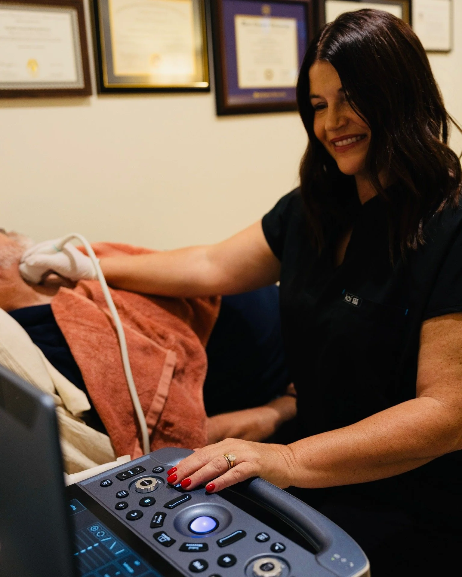 A smiling woman with dark hair is meditating a patient's arm while working on medical equipment in a clinic or hospital setting. The patient is lying on a bed covered with an orange towel, and the woman is wearing a black uniform with a name tag. Several framed certificates or diplomas hang on the beige wall behind them.