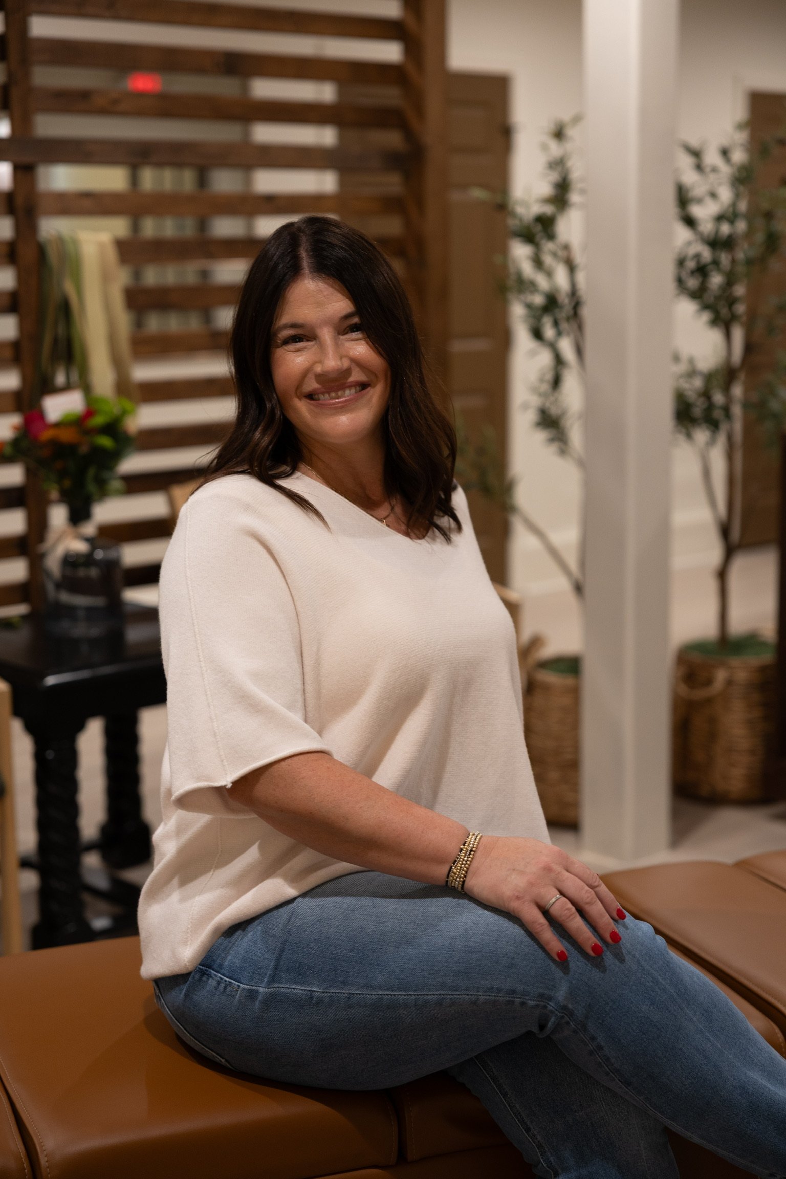 A woman with brown hair, smiling and wearing a white top and blue jeans, sitting on a brown leather seat indoor with wooden and plant decor in the background.