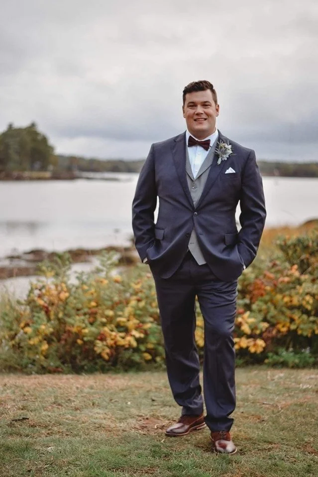 A young man in a dark suit and bow tie standing outdoors by a body of water with autumn foliage in the background.