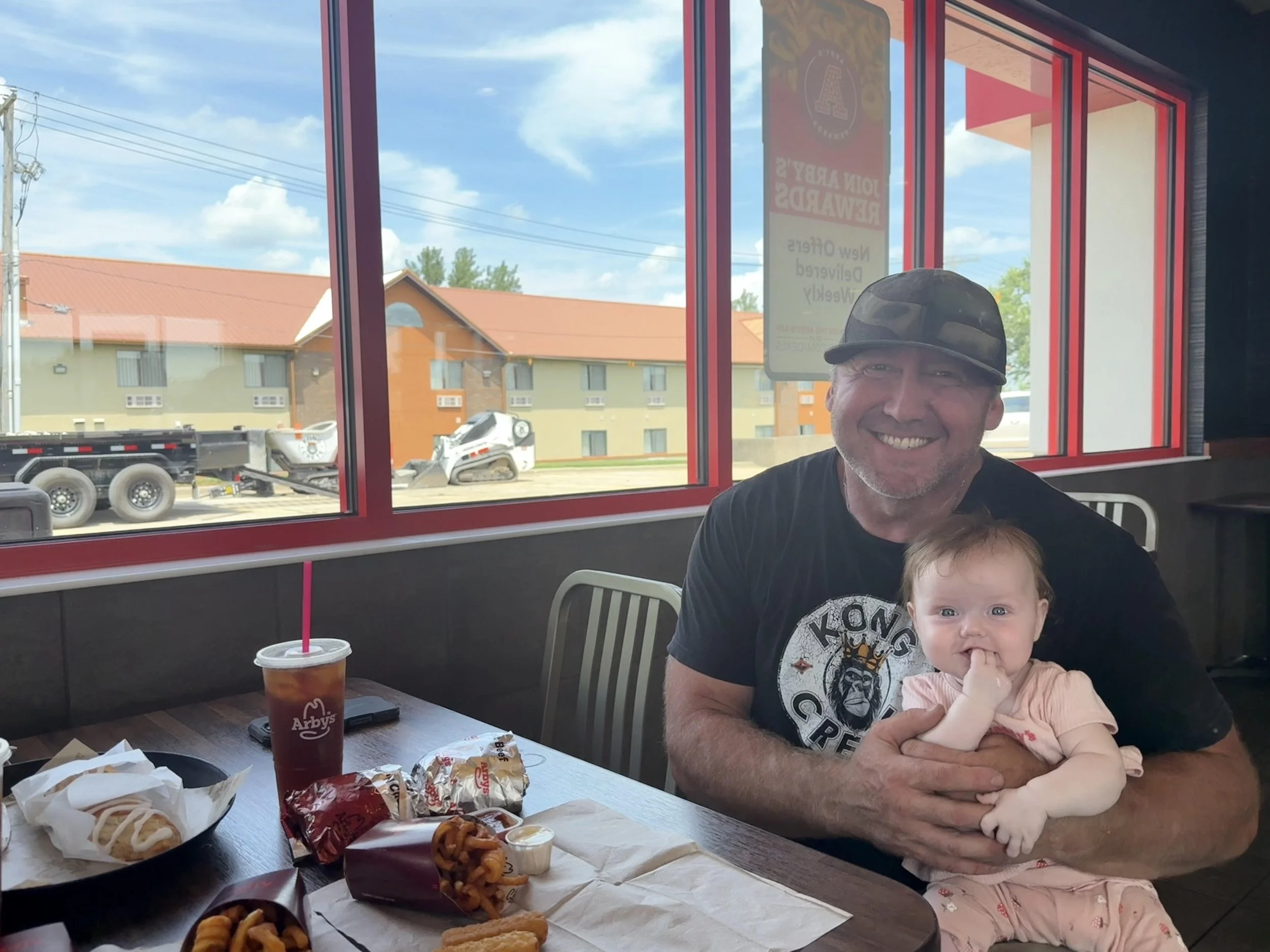 A man and a baby smiling and sitting at a table inside Arby's restaurant. The table has empty food trays, curly fries, and an iced drink. Outside, buildings and a truck are visible through large windows.