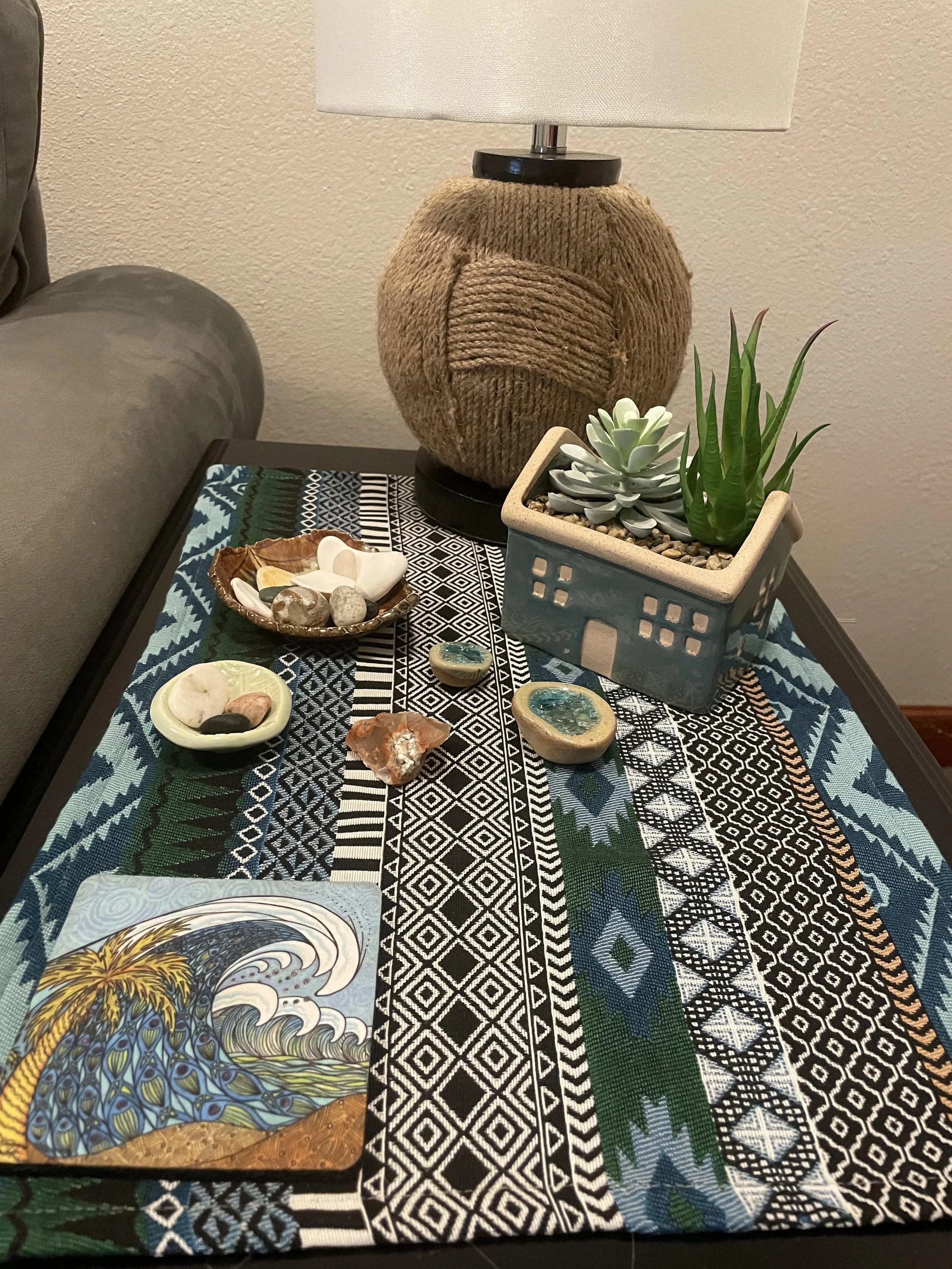 Side table with handmade decorative items, including a ceramic dish of shells and stones, a small plate of stones and shell, and a tray with artwork of a peacock, along with a lamp with a rope-wrapped base, a potted succulent and cactus, and a patterned table runner.