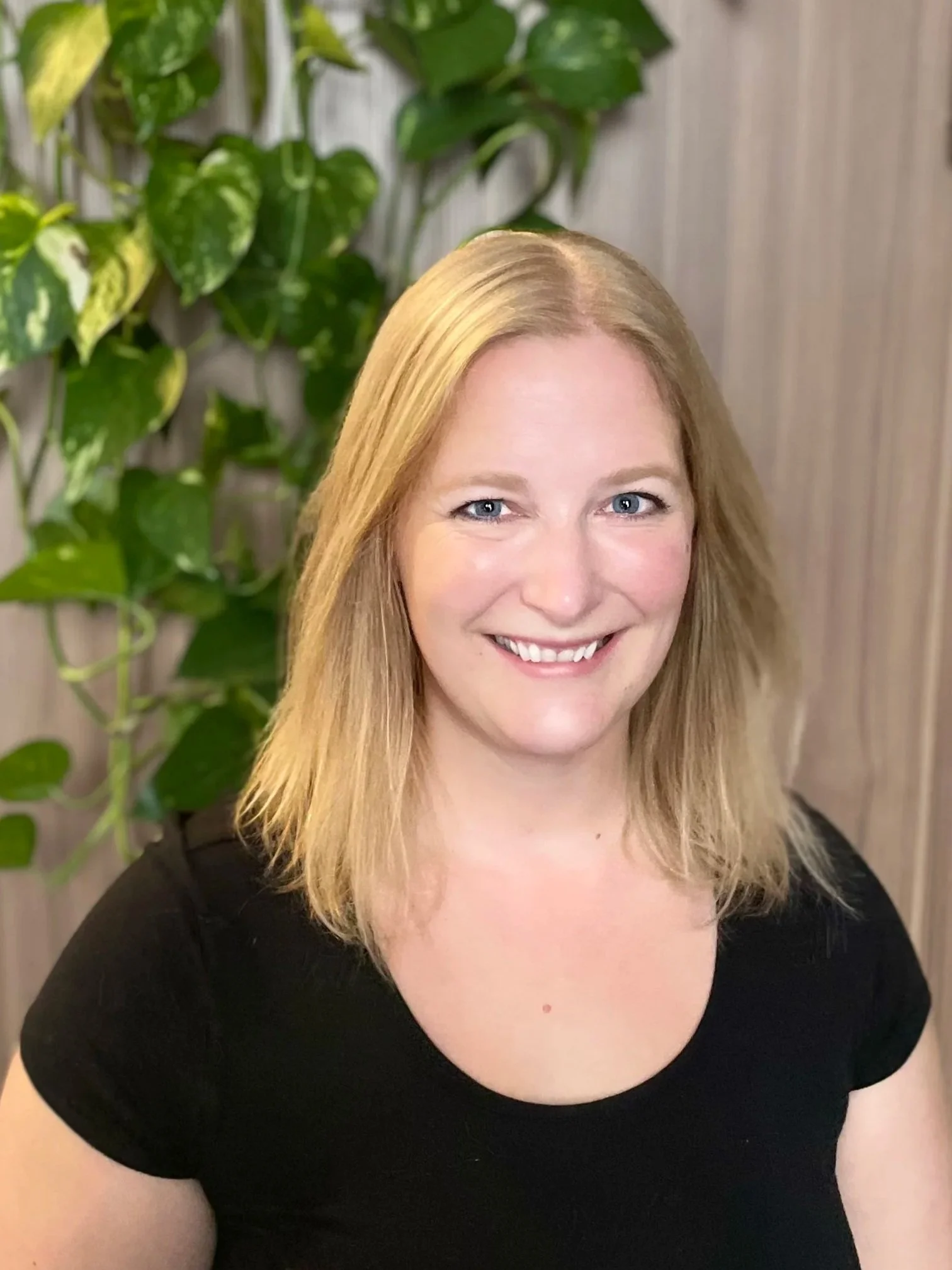 A woman with blonde hair smiling, wearing a black top, standing in front of green leafy plants and a light-colored wall.