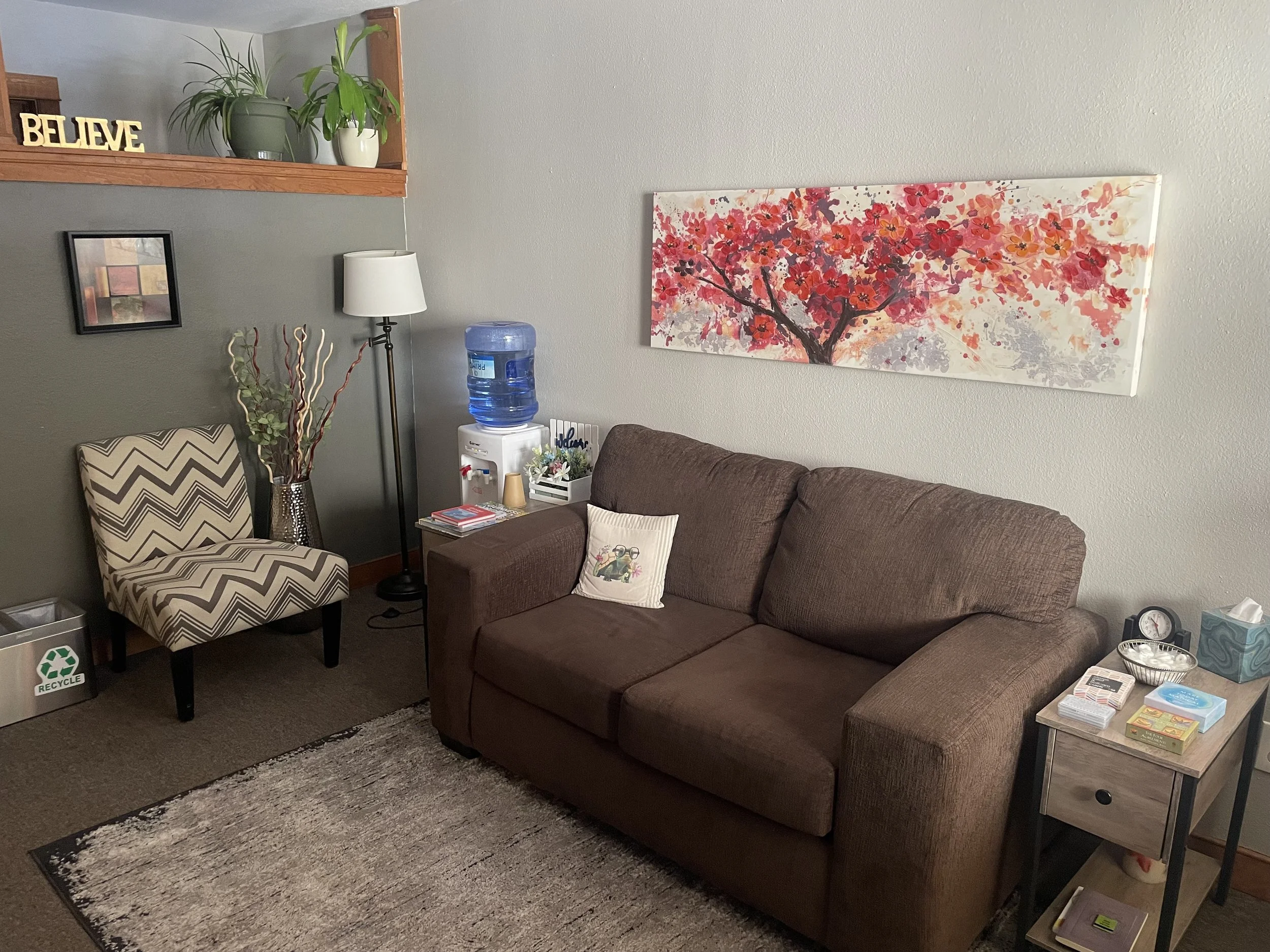 A living room with a brown sofa, a table with books and tissues, a patterned armchair, a floor lamp, a water dispenser, and a painting of a flowering tree on the wall.