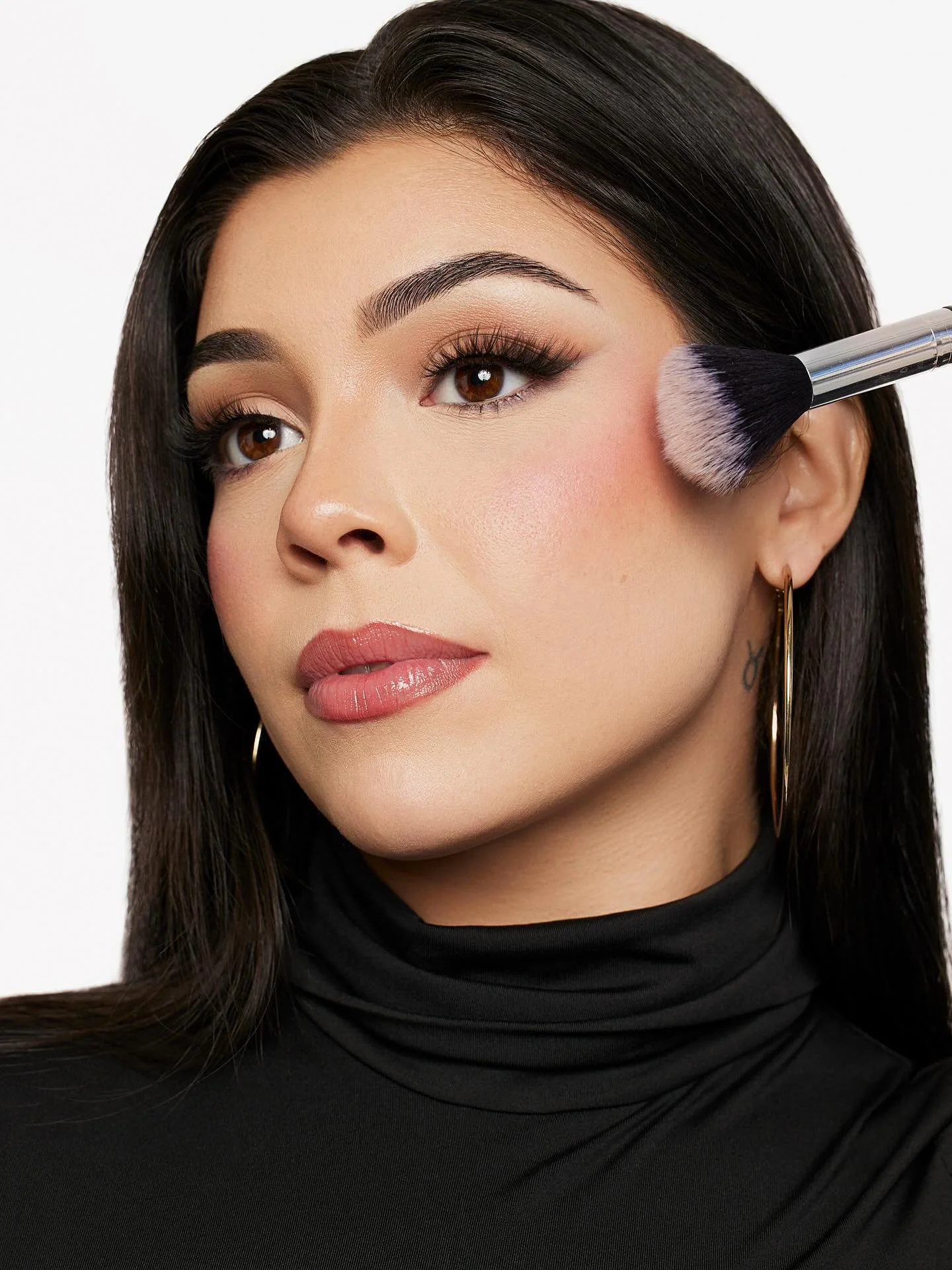 Andrea Salomon with long dark hair receiving makeup application using a fluffy brush on her cheek, wearing a black turtleneck and gold hoop earrings.