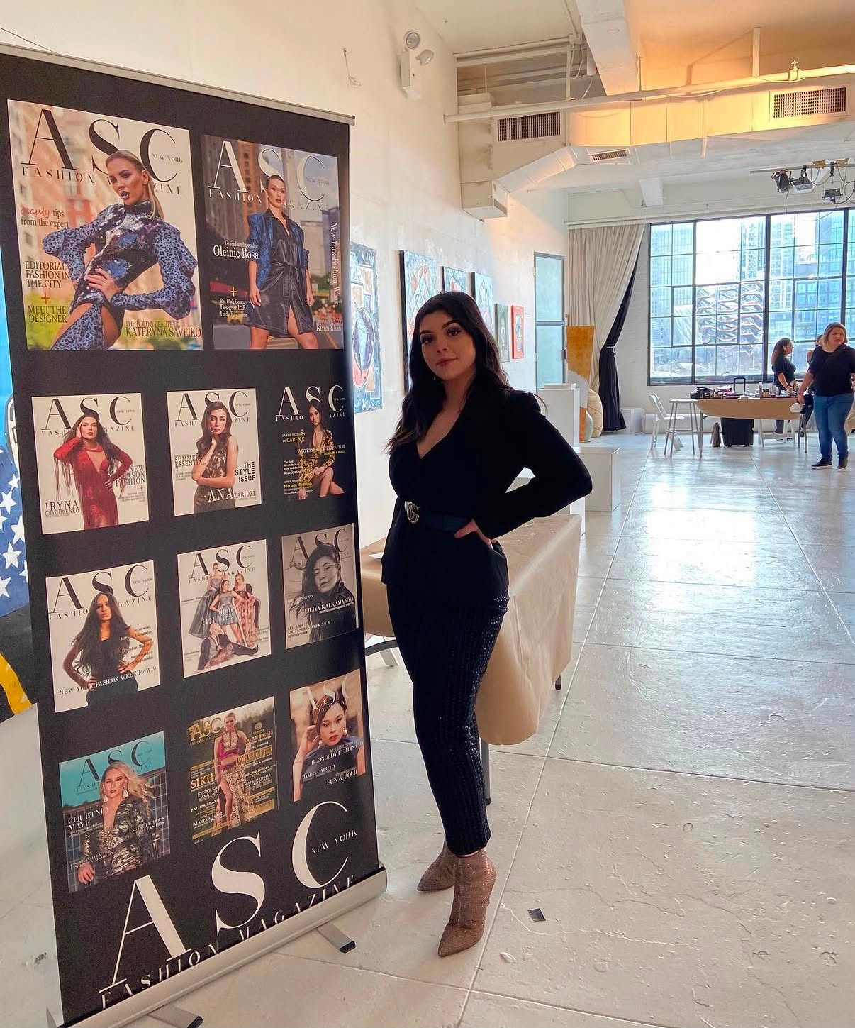Andrea Salomon standing next to a display board featuring multiple magazine covers. Set in an art gallery or event space with large windows, artwork on the walls, and a table with items in the background.