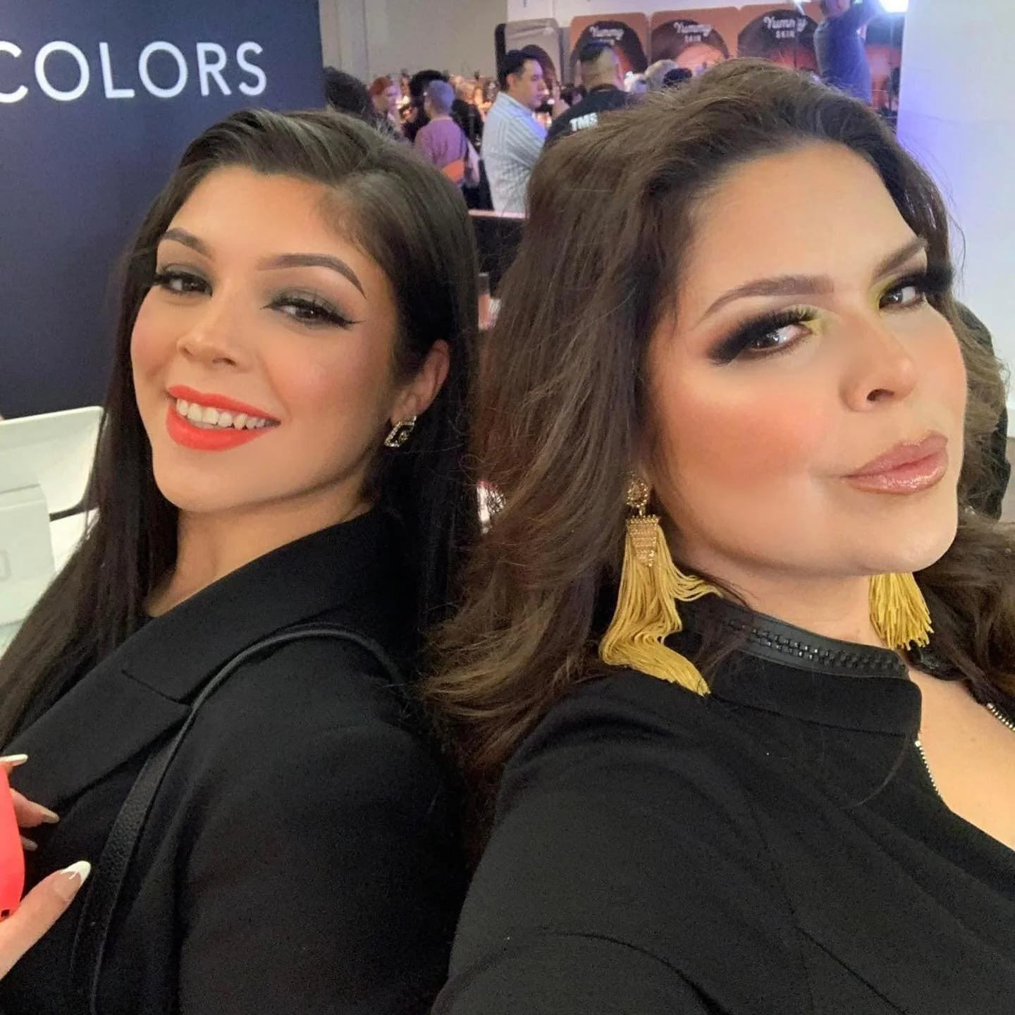 Andrea Salomon and Brenda Rodriguez posing at NYFW. Andrea has red lipstick and a black outfit, while Brenda has yellow eye makeup, blonde earrings, and a black jacket with yellow fringe details.