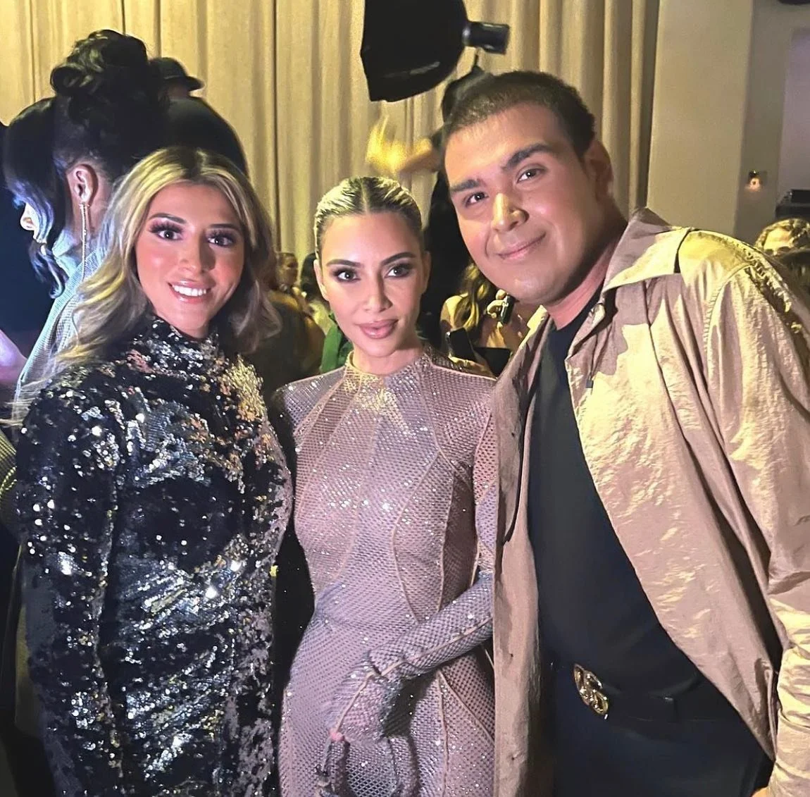 Kim Kardashian and two more people dressed in glamorous outfits with a background of curtains and other attendees.