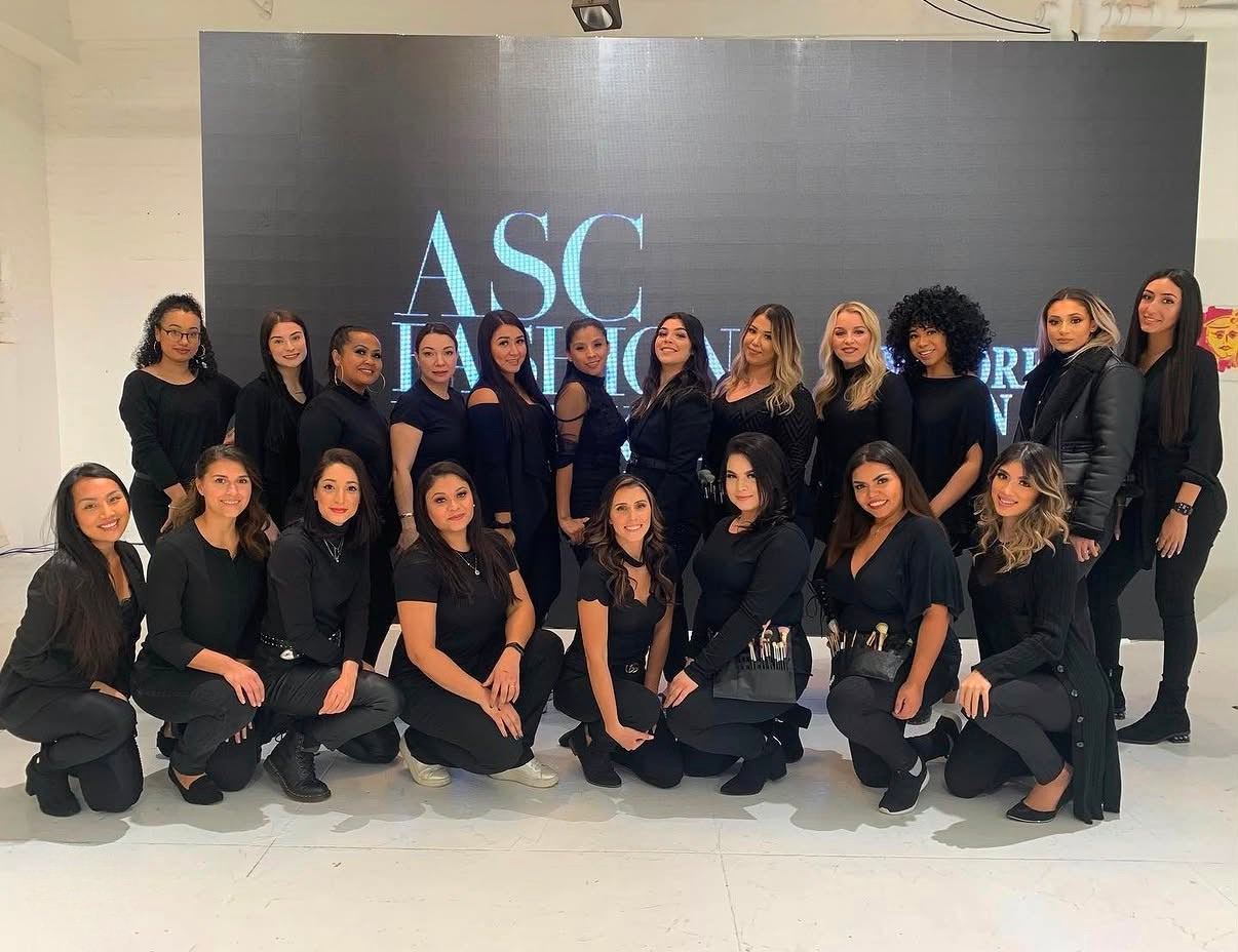 Group of women in black clothing standing and kneeling in front of a large digital screen displaying the text 'ASC'. Some women are wearing makeup and holding makeup tools. Andrea Salomon is in the group.