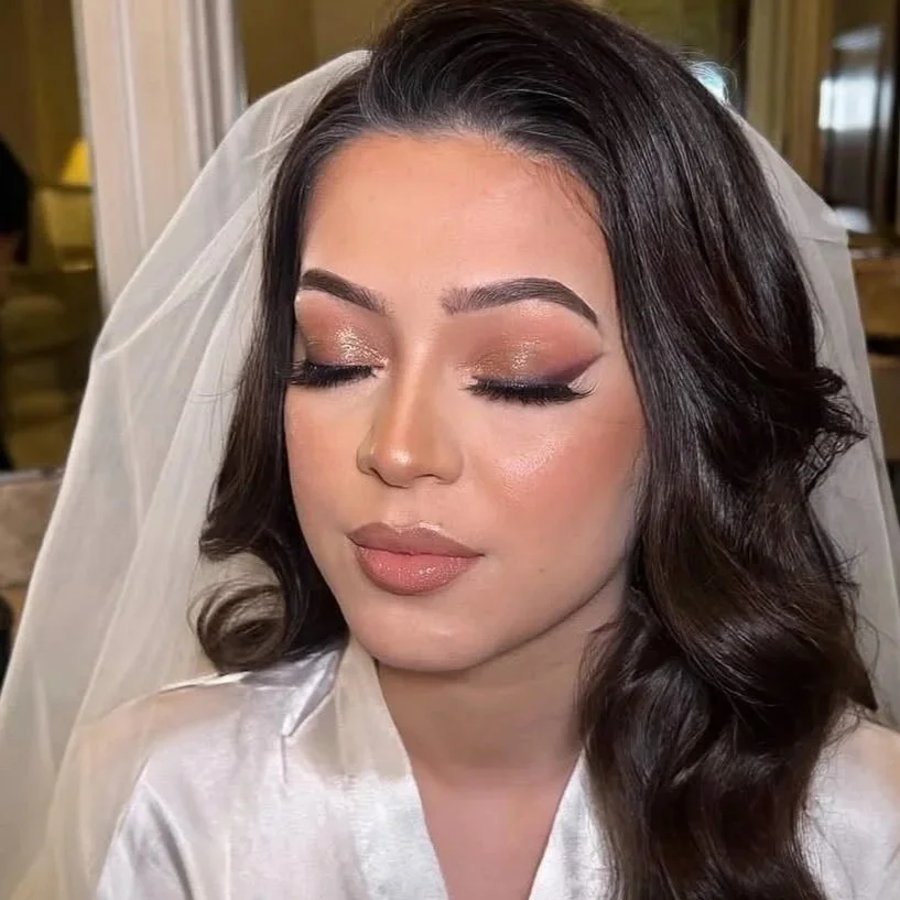 A bride with dark brown hair styled in loose waves, wearing bridal makeup, a white silk robe, and a white veil. Her eyes are closed, showcasing shimmery eyeshadow and long eyelashes.