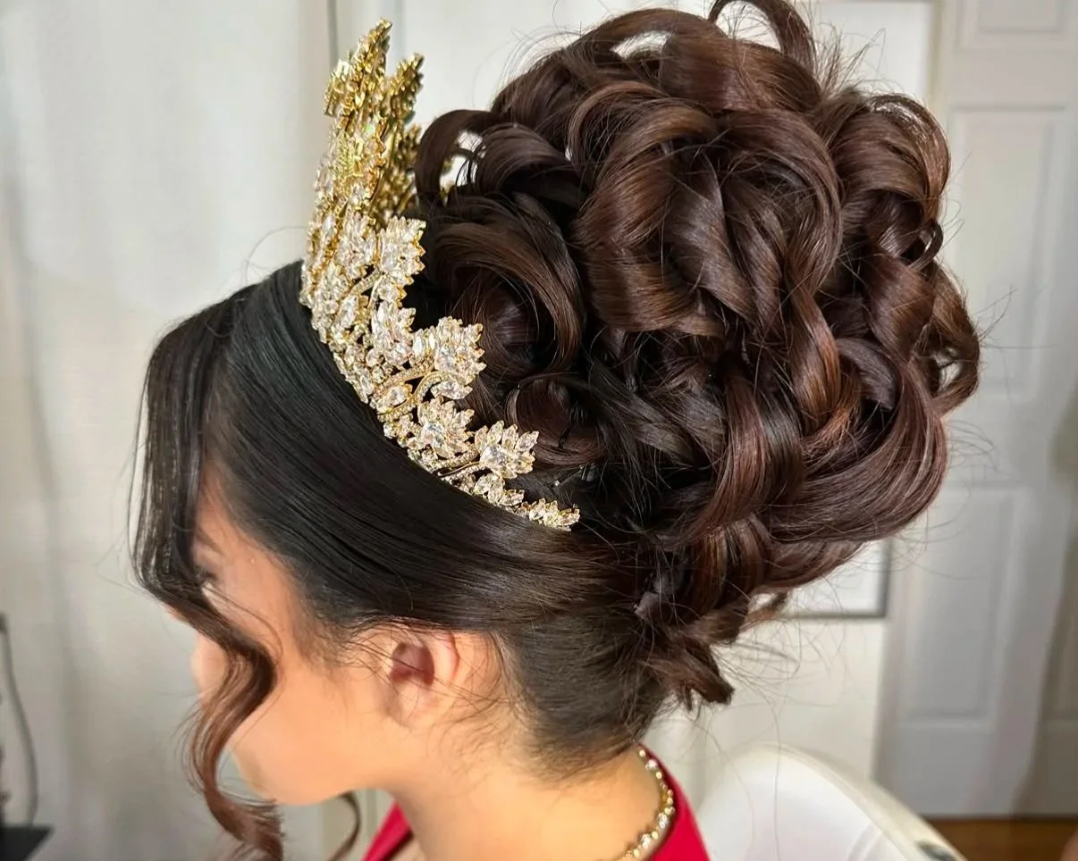 Elegant updo hairstyle with large curls and a decorative gold and rhinestone tiara, worn by a woman with dark hair and side-swept bangs, in a bridal or formal setting.