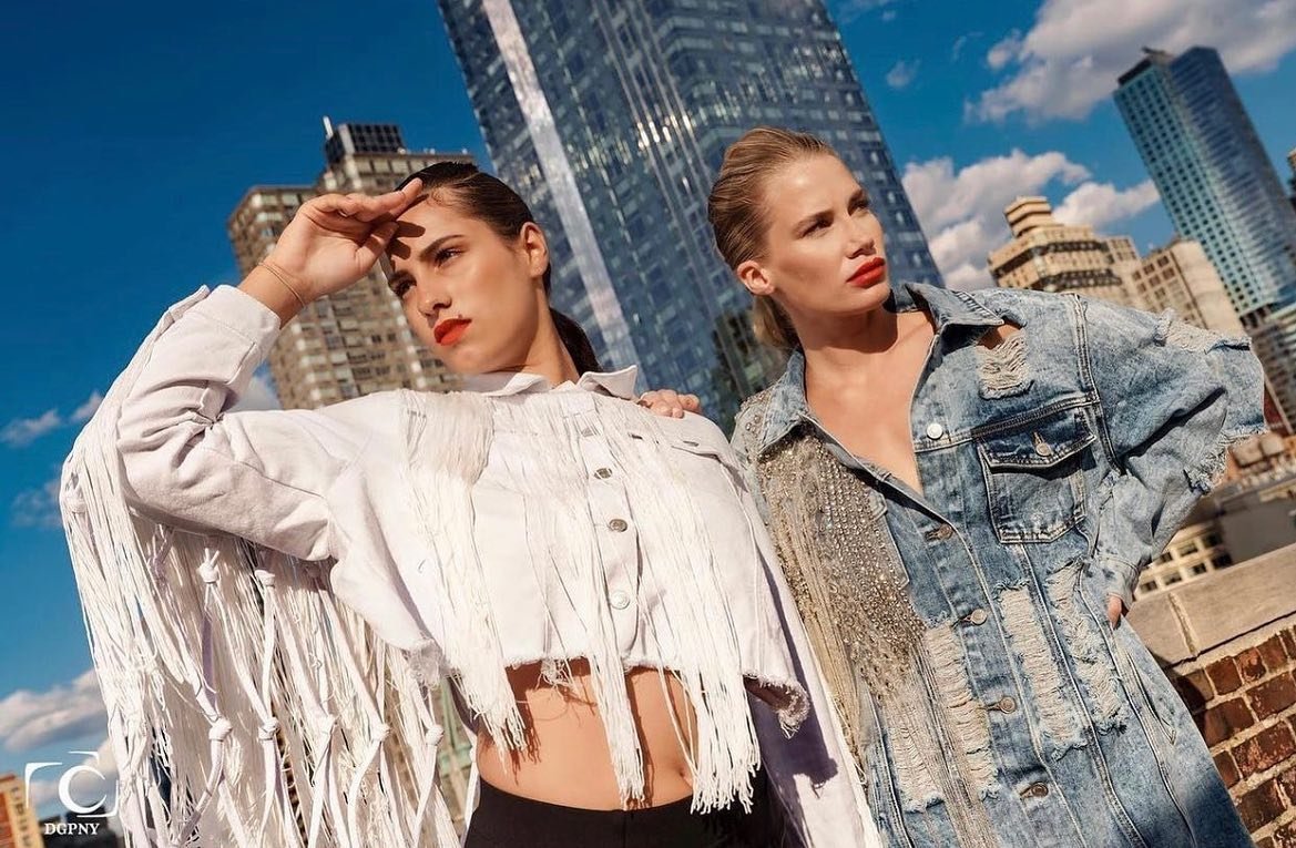 Two women standing outdoors in an urban setting with skyscrapers behind them, one with dark hair and the other with blonde hair, both wearing edgy denim jackets and posing confidently for a fashion magazine
