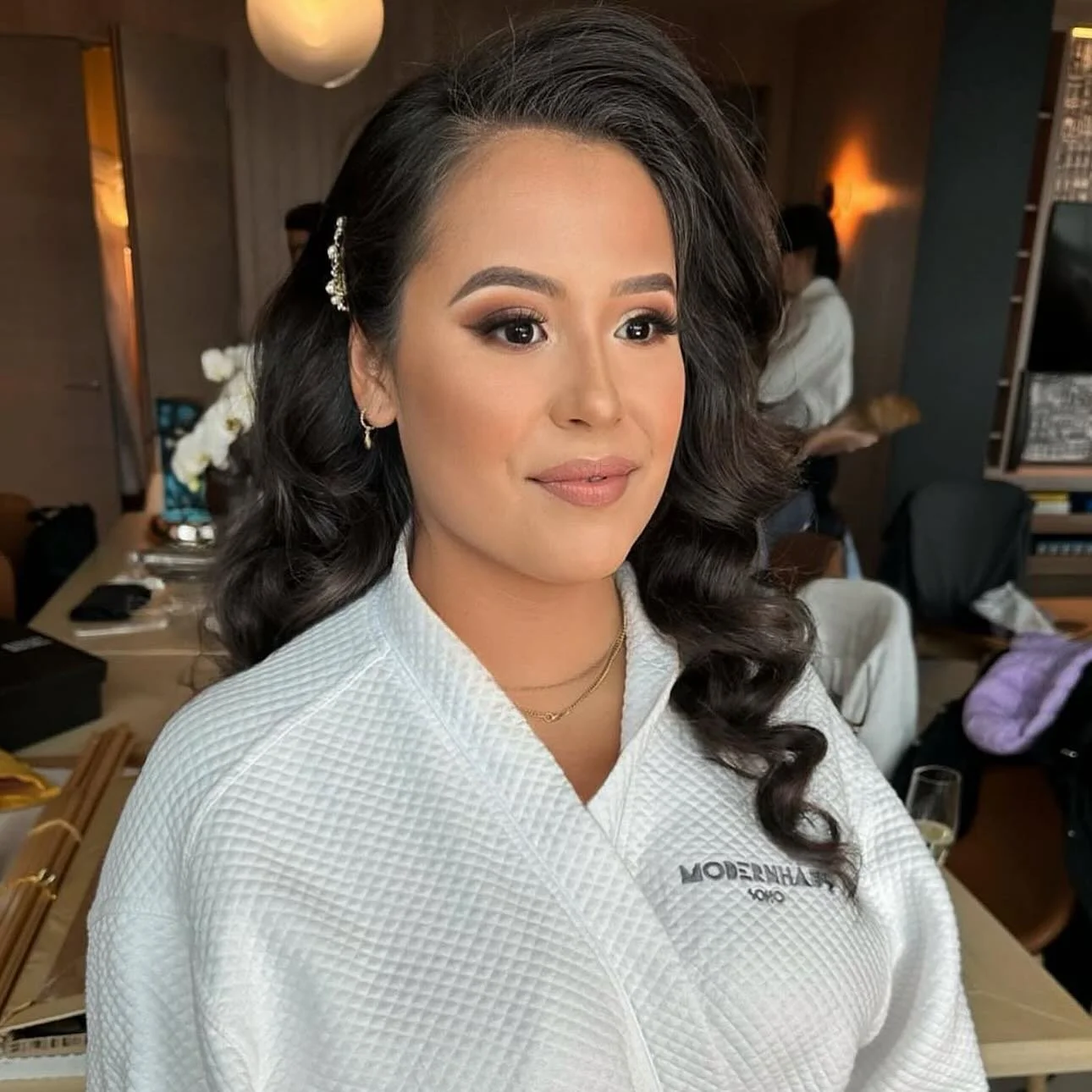 A bride with dark, styled hair and bridal makeup, wearing a white textured jacket, in a bridal style prep room.