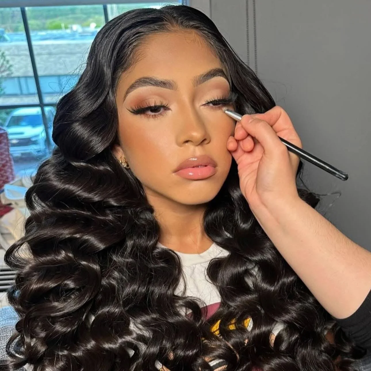A woman with long, dark, curly hair gets her makeup done by makeup artist Andrea Salomon applying eyeshadow with a brush in a well-lit room near a window.