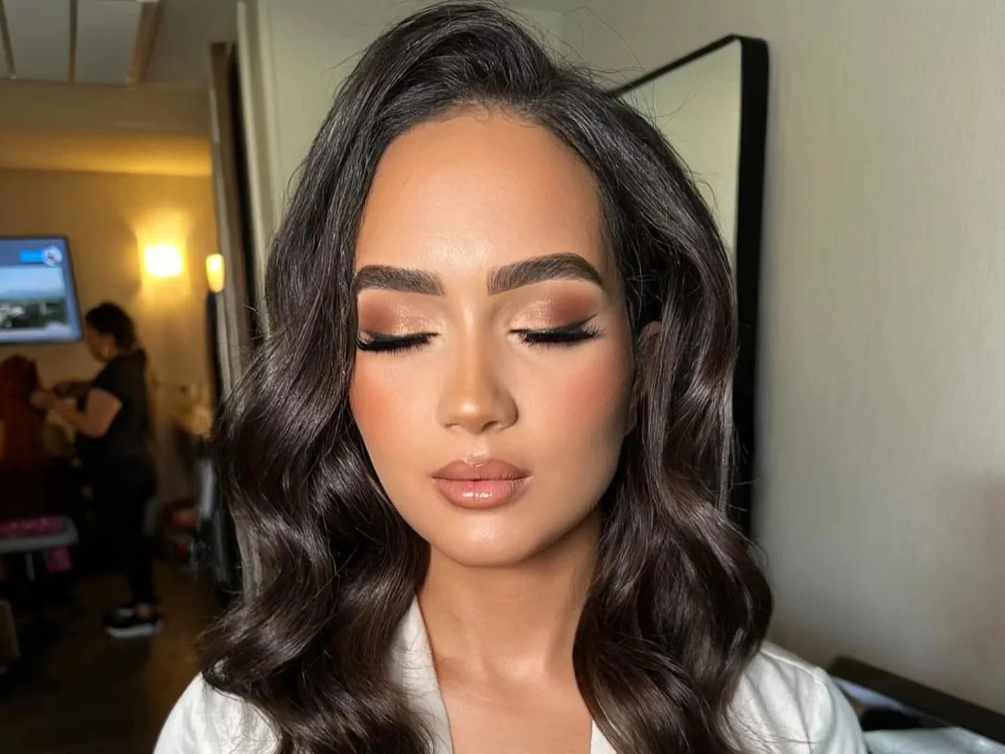 Close-up of a bride with long, wavy dark hair, closed eyes, and bridal makeup including eyeshadow, eyeliner, and lipstick, in a room with a mirror and a person in the background.