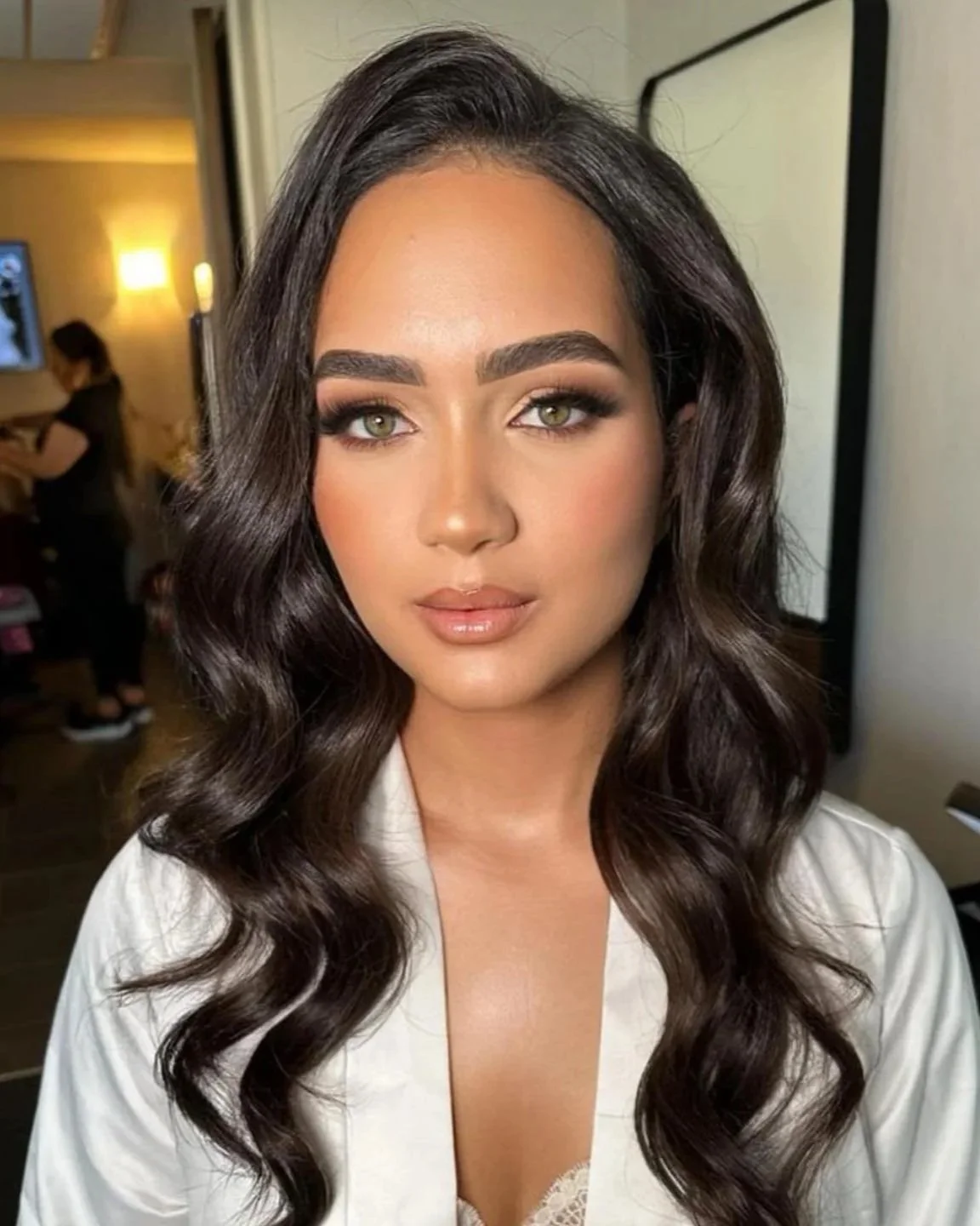 Close-up of a woman with long, wavy dark hair, makeup, and a white robe, in a room with a mirror and a person in the background.