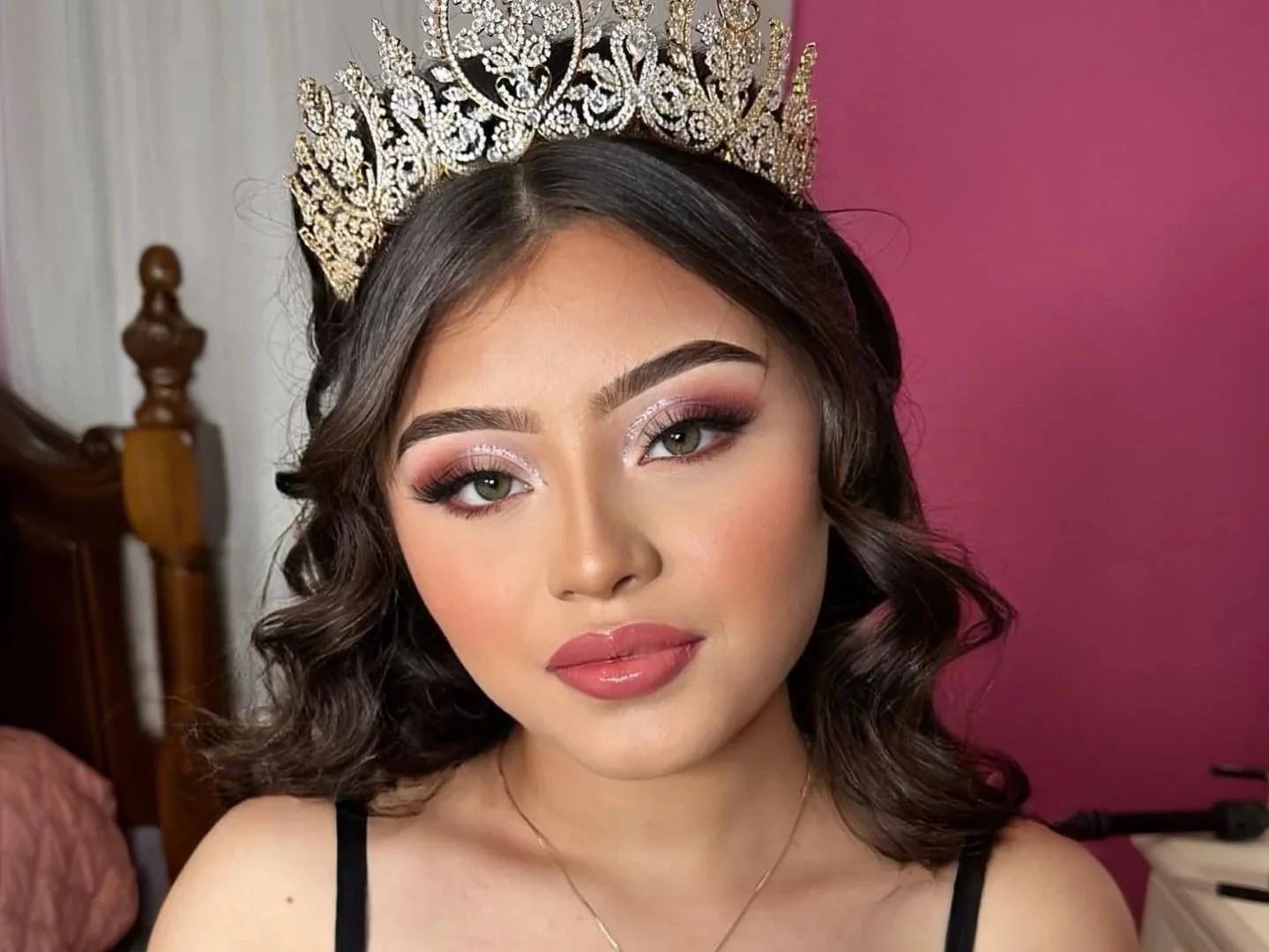 A woman wearing a large ornate tiara, with makeup and wavy shoulder-length hair, posing indoors with a pink wall background.