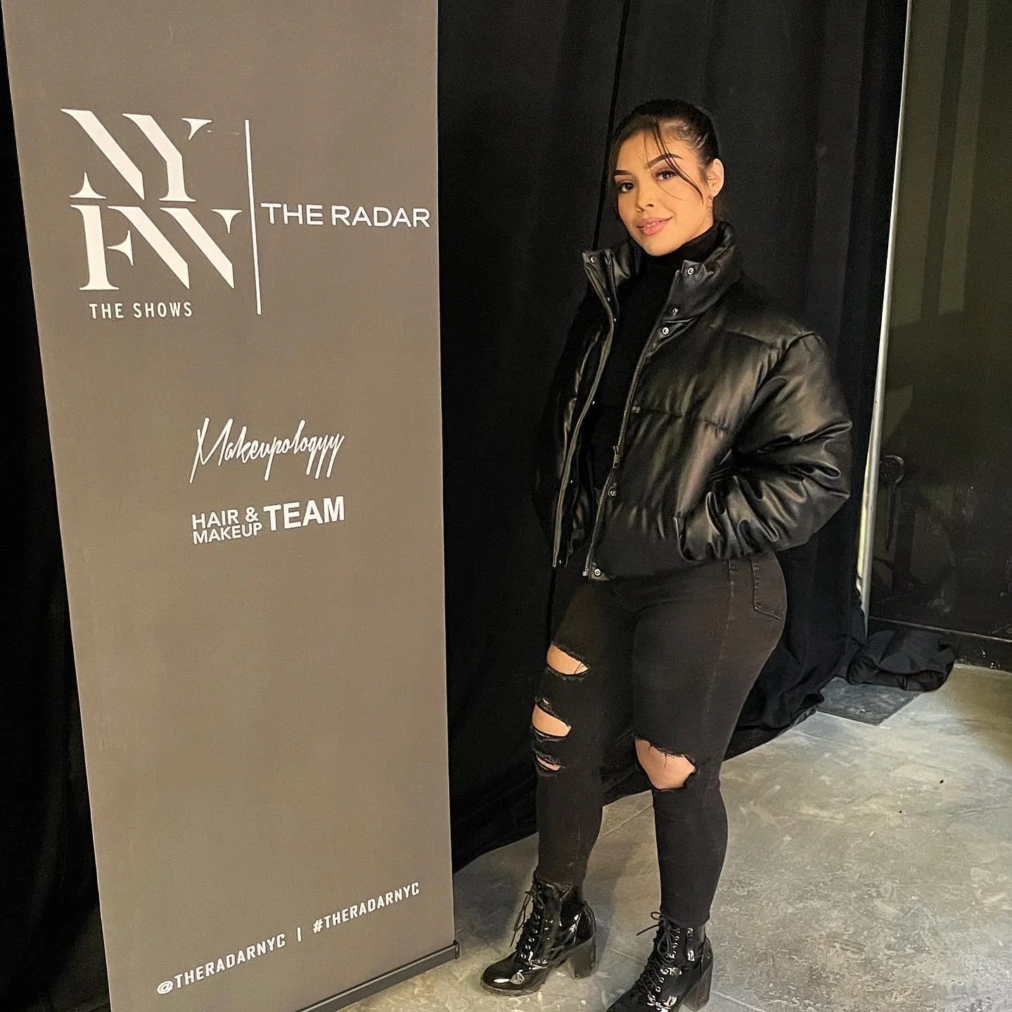 Andrea Salomon standing next to a promotional banner for New York Fashion Week and makeup team, wearing a black leather jacket, ripped black jeans, and black lace-up ankle boots.