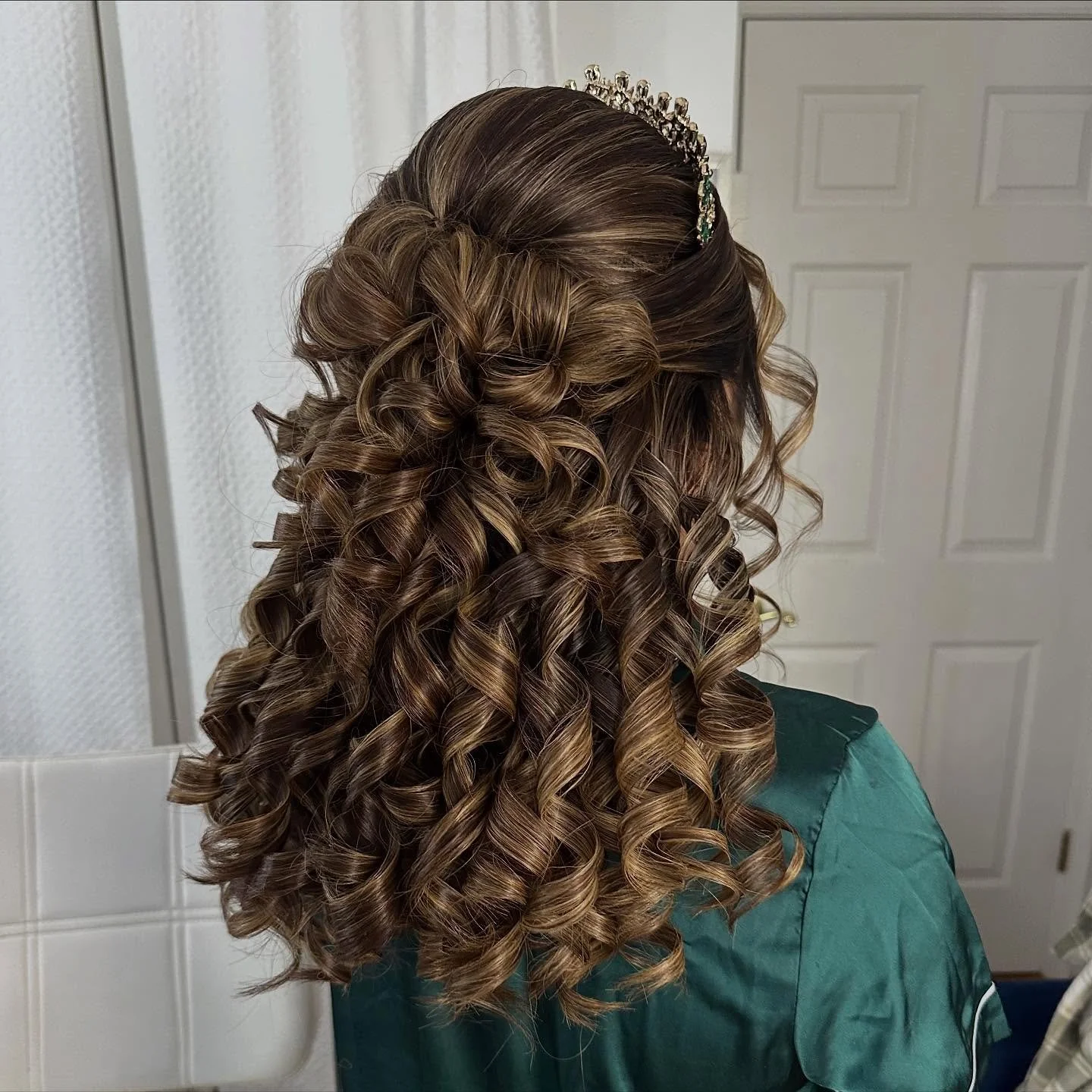 Back view of a woman with long, dark blonde curly hair styled with volume, decorated with a silver tiara with green accents, wearing a teal gown.