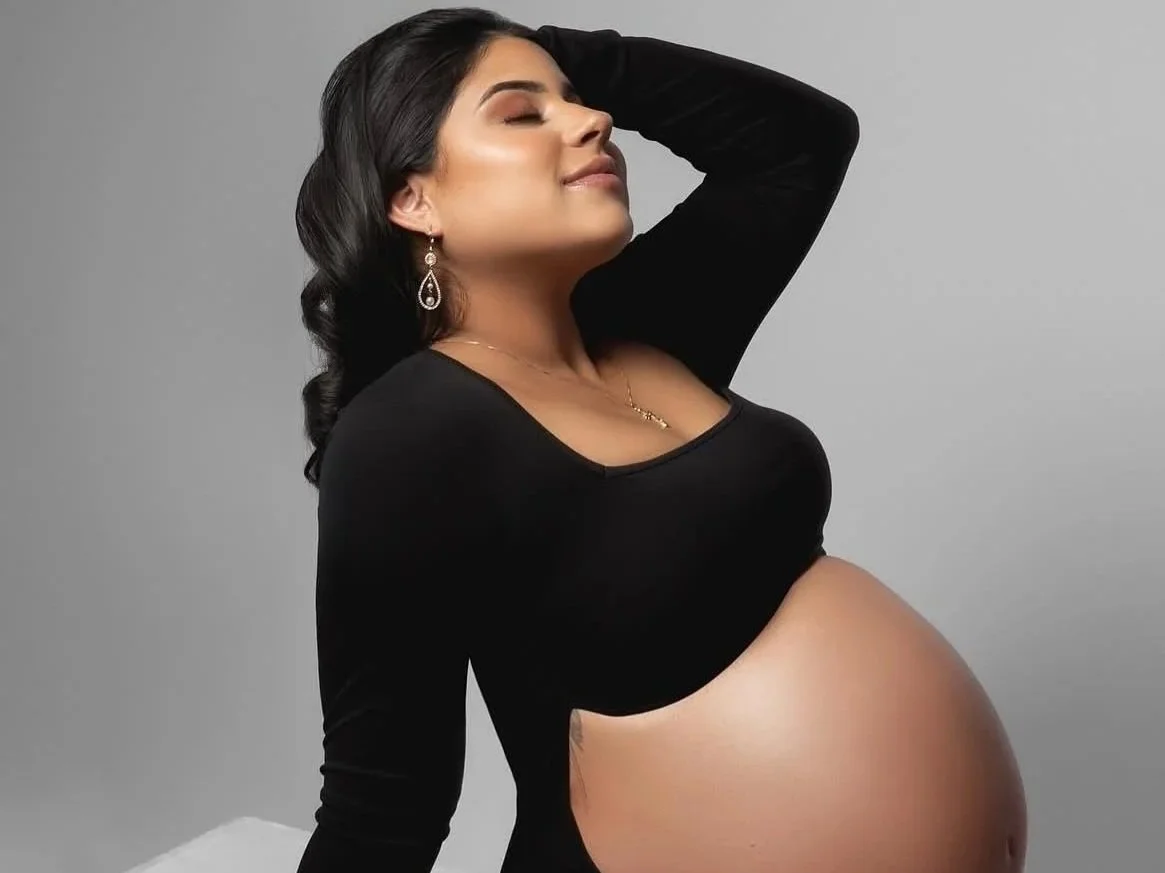 Pregnant woman posing for a photoshoot with professionally done makeup with dark hair, wearing a black top, earrings, and jewelry.