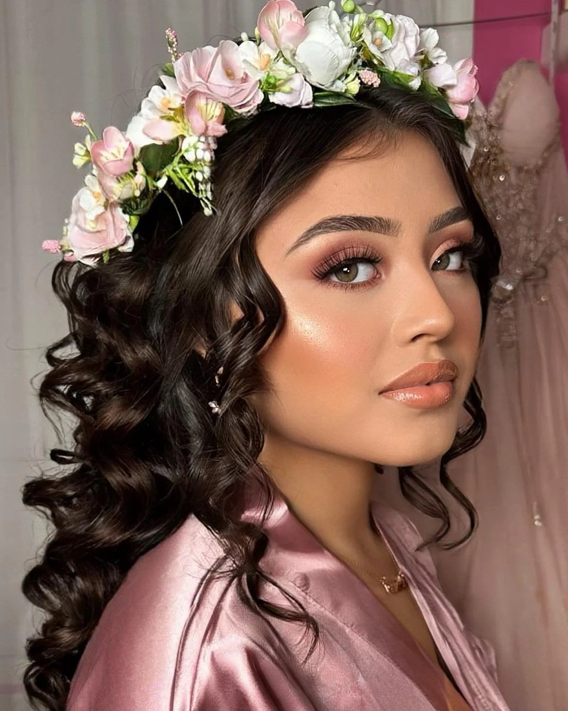 A young woman with dark, curly hair wearing a pink satin robe and a floral crown, with makeup including foundation, blush, highlighter, shimmery eye makeup, and nude lipstick.