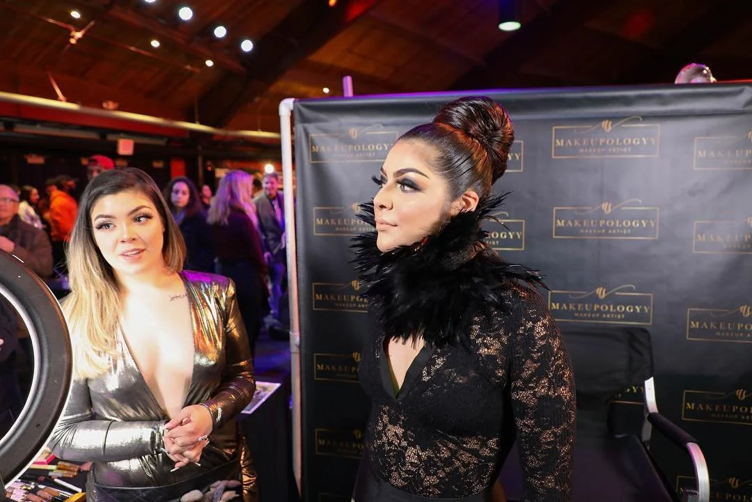 A woman with styled hair and makeup posing at a makeup booth with a lace outfit and feathered collar, while Andrea Salomon has loose hair and golden metallic outfit talks nearby at the makeup event.