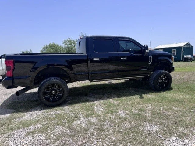 Black pickup truck with large tires parked on gravel, with a small house and trees in the background.