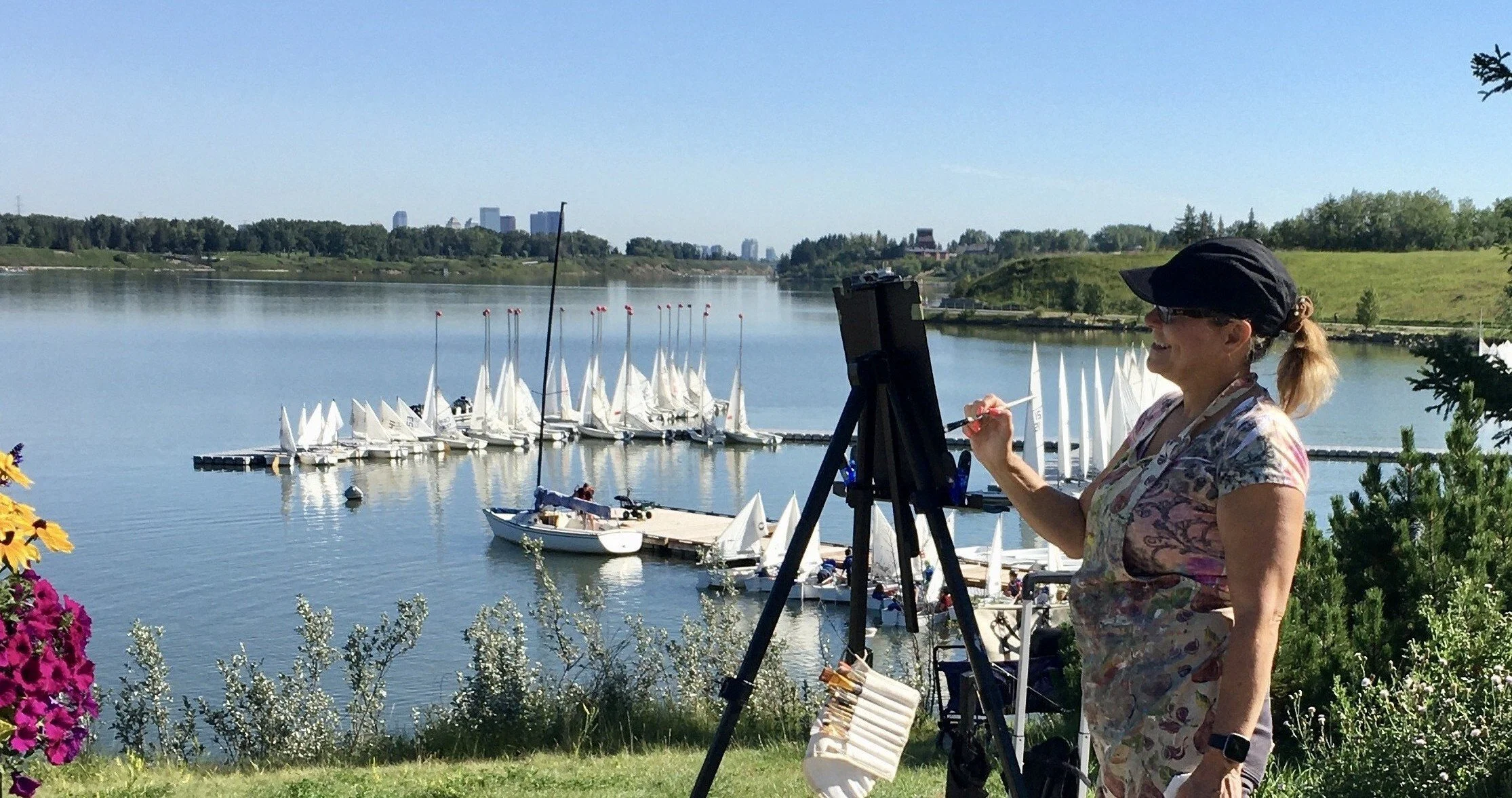 A woman painting outdoors near a lake with sailboats docked, flowers and greenery in the foreground, and city buildings in the background.