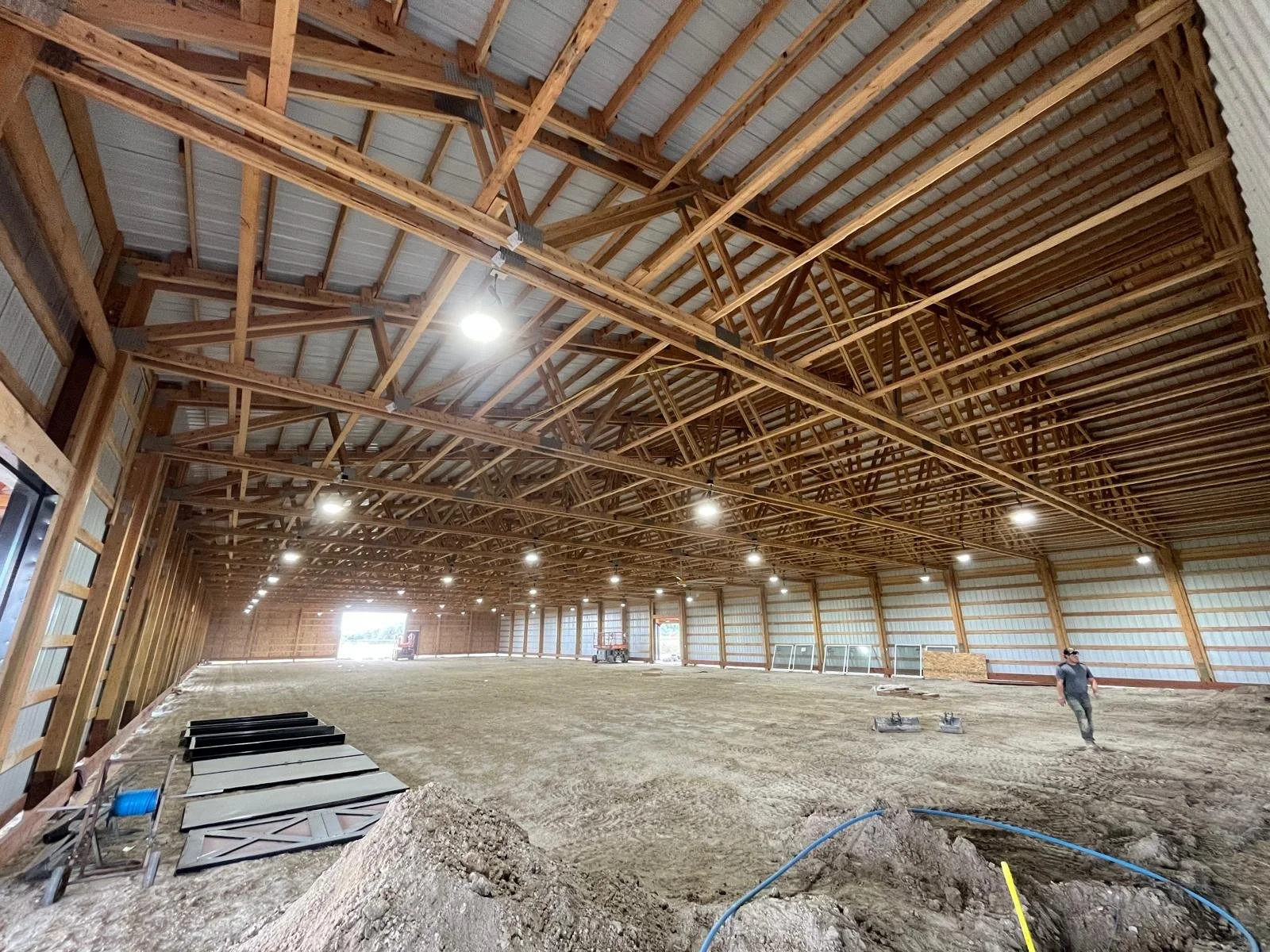 Interior of a large unfinished barn or warehouse with exposed wooden truss roof, metal siding walls, and dirt floor, with construction materials and workers present.