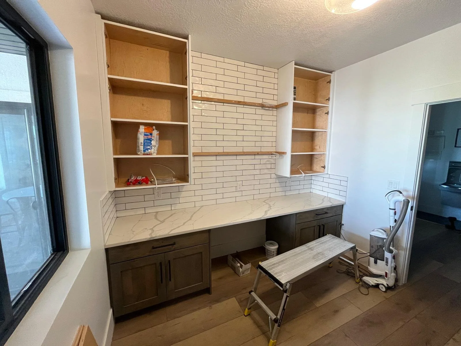 Kitchen renovation in progress with open upper cabinets, white subway tile backsplash, marble countertop, and a vacuum cleaner on the right side.
